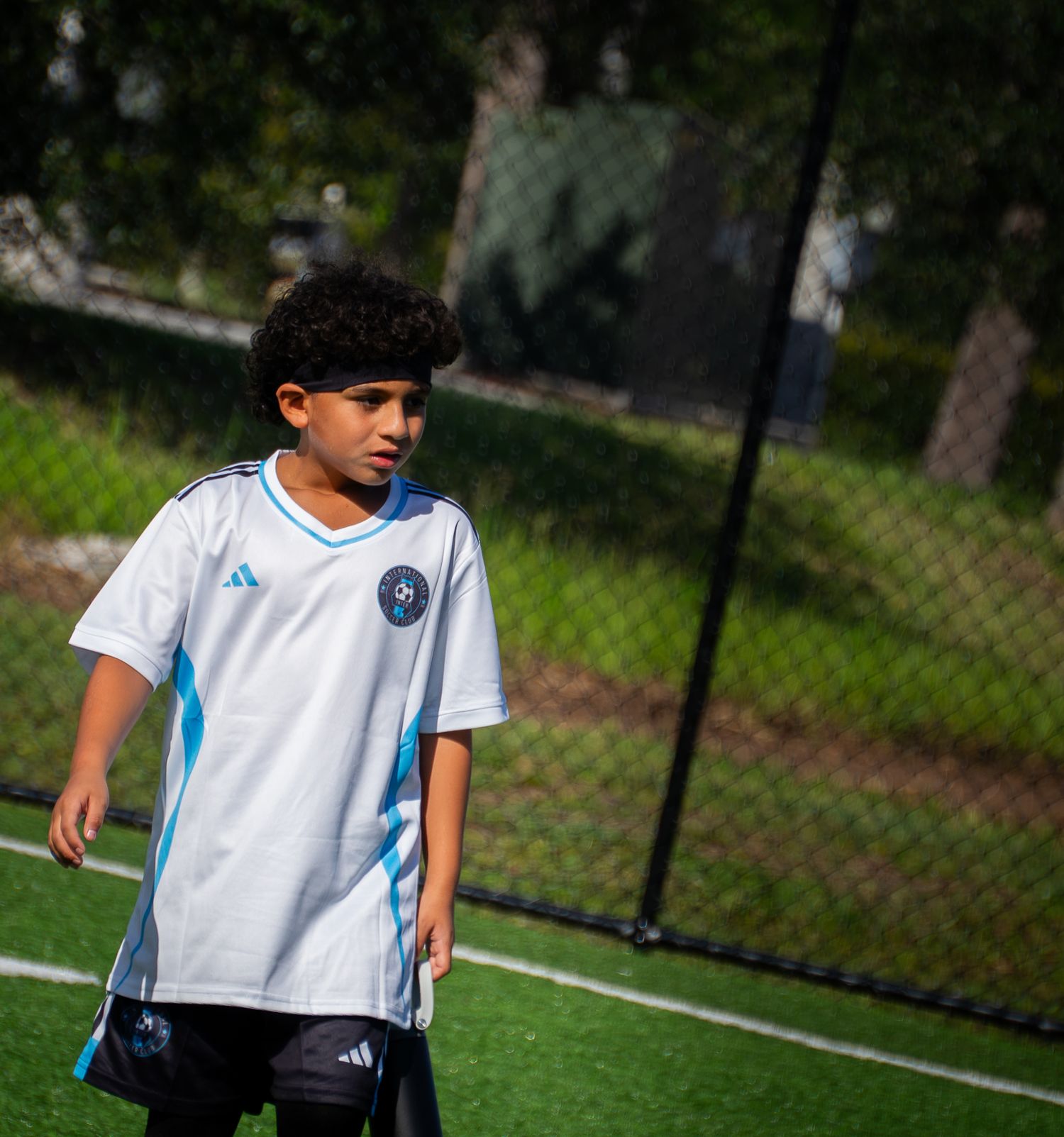 Young soccer player in white and blue Adidas jersey walks on green turf near a chain-link fence.