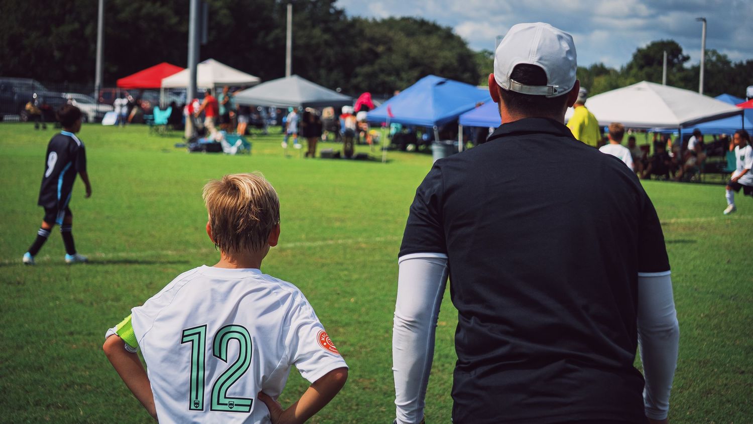 Youth soccer player wearing number 12 jersey stands beside adult coach watching game at tournament.
