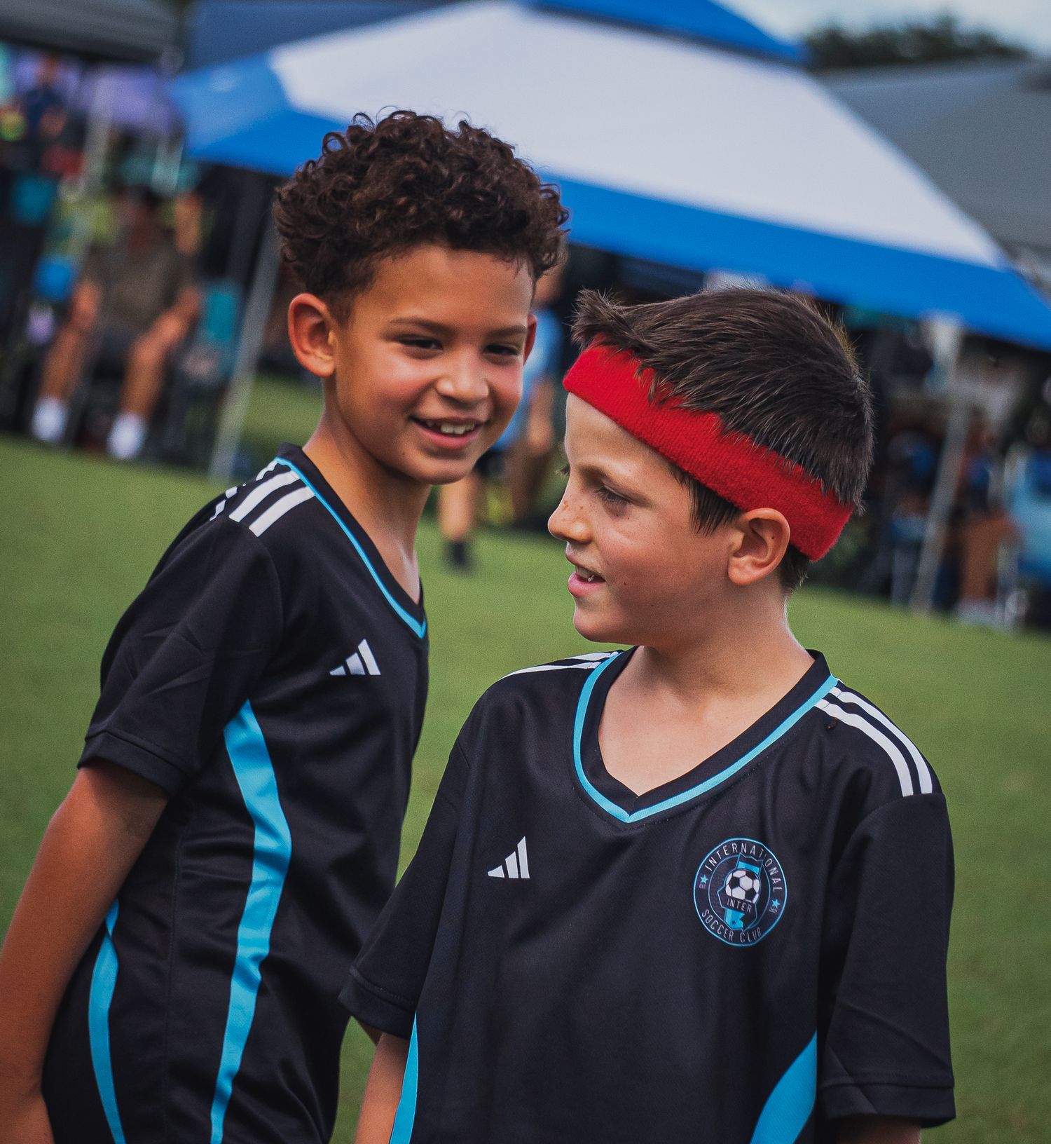 Two young boys in black Adidas soccer uniforms smile together on field at youth tournament.