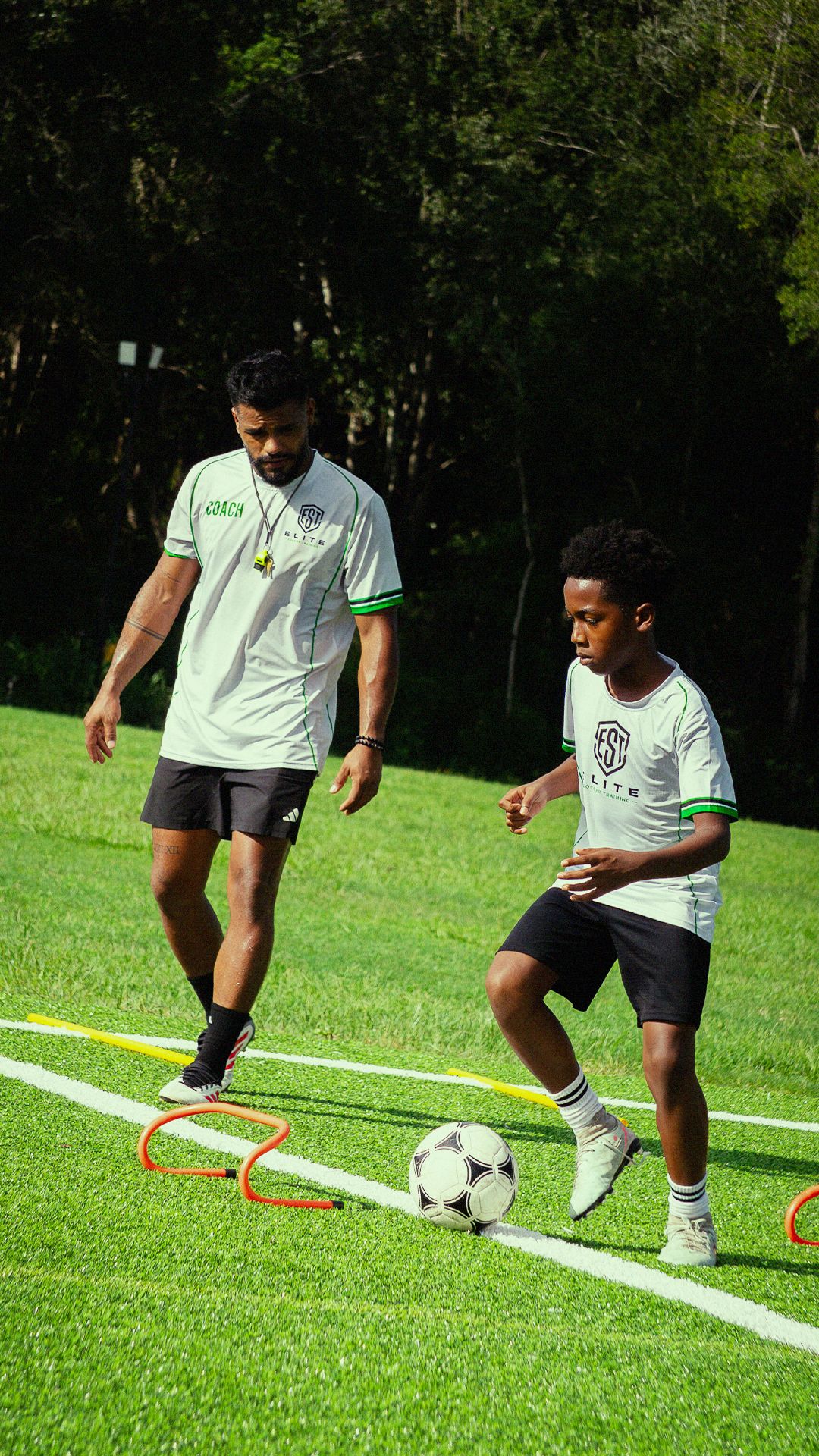 Soccer coach in white jersey guides young player through dribbling drills on a green turf field.