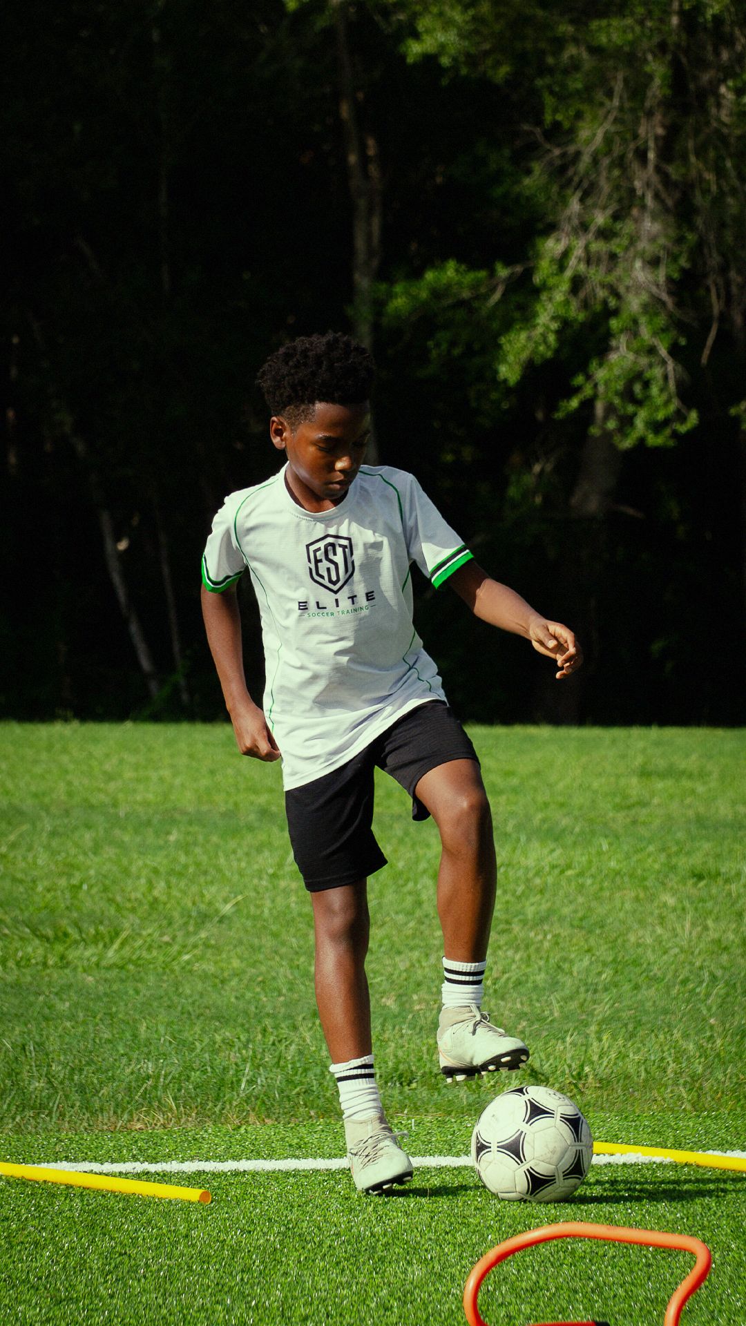 Young soccer player in white Elite jersey practices dribbling with a soccer ball on a green turf field.