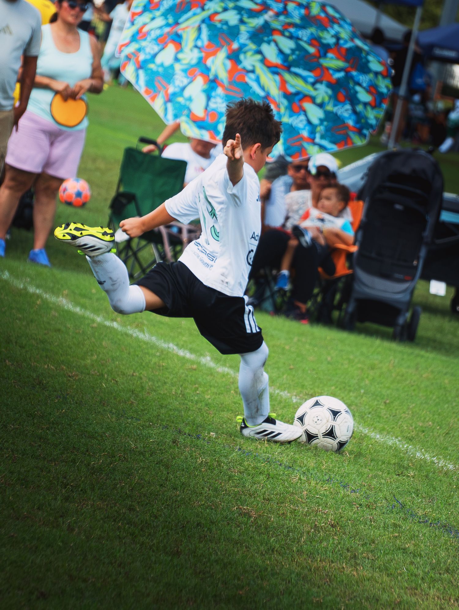 Youth soccer player in white kit executing a powerful kick during an outdoor grass field match.