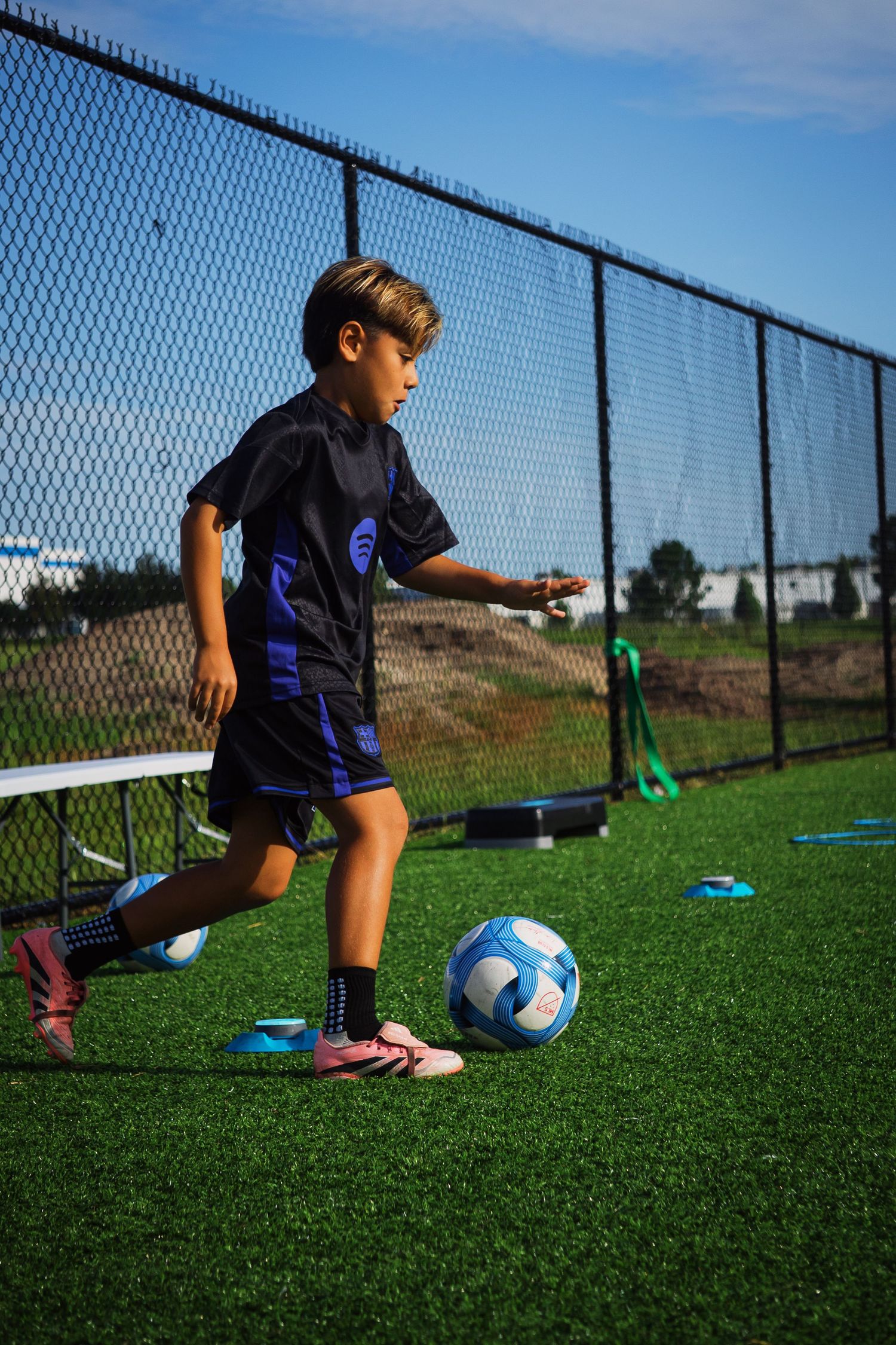 Young boy in dark blue soccer uniform practices dribbling a blue and white ball on turf field.