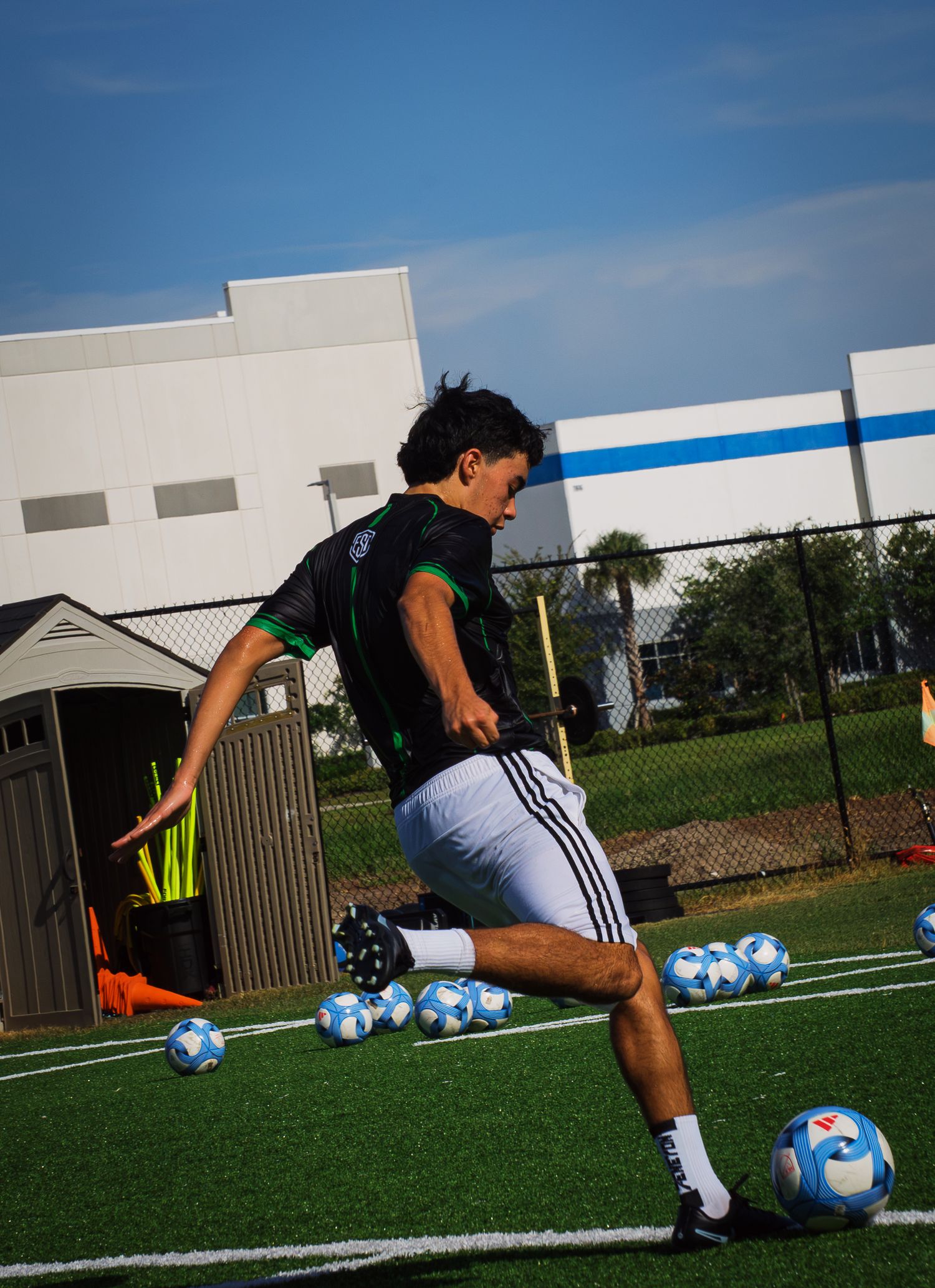 Teen soccer player in black jersey kicking a ball on turf field surrounded by multiple soccer balls.