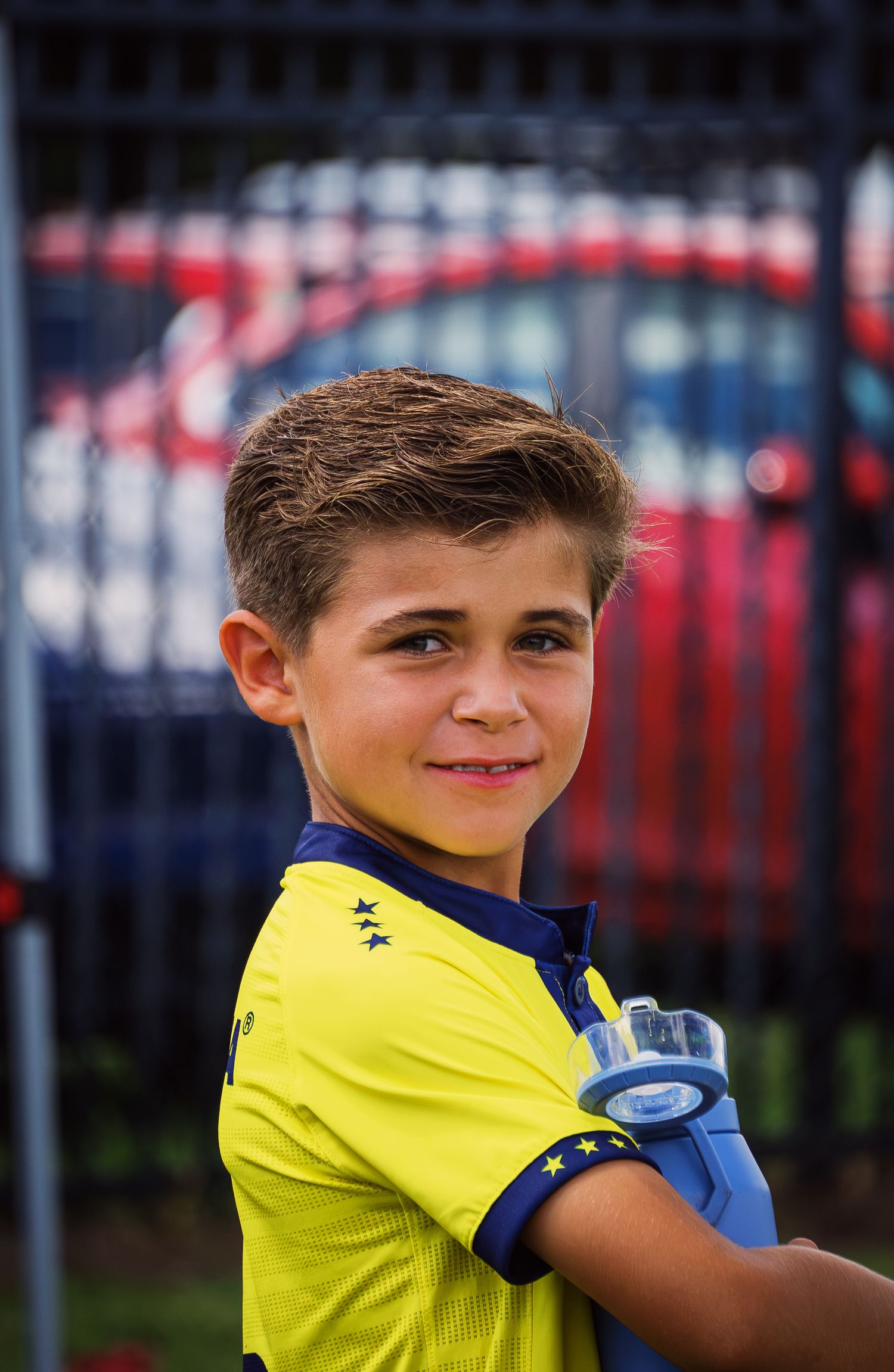 Smiling young boy in yellow and blue soccer jersey holds water bottle at outdoor field.