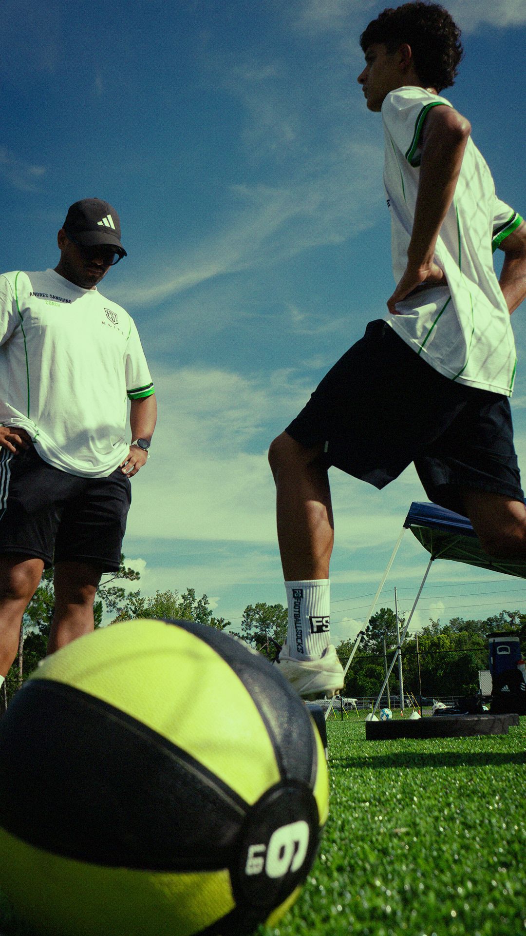 Two youth soccer players in white Adidas jerseys train on a green field with a medicine ball in the foreground.