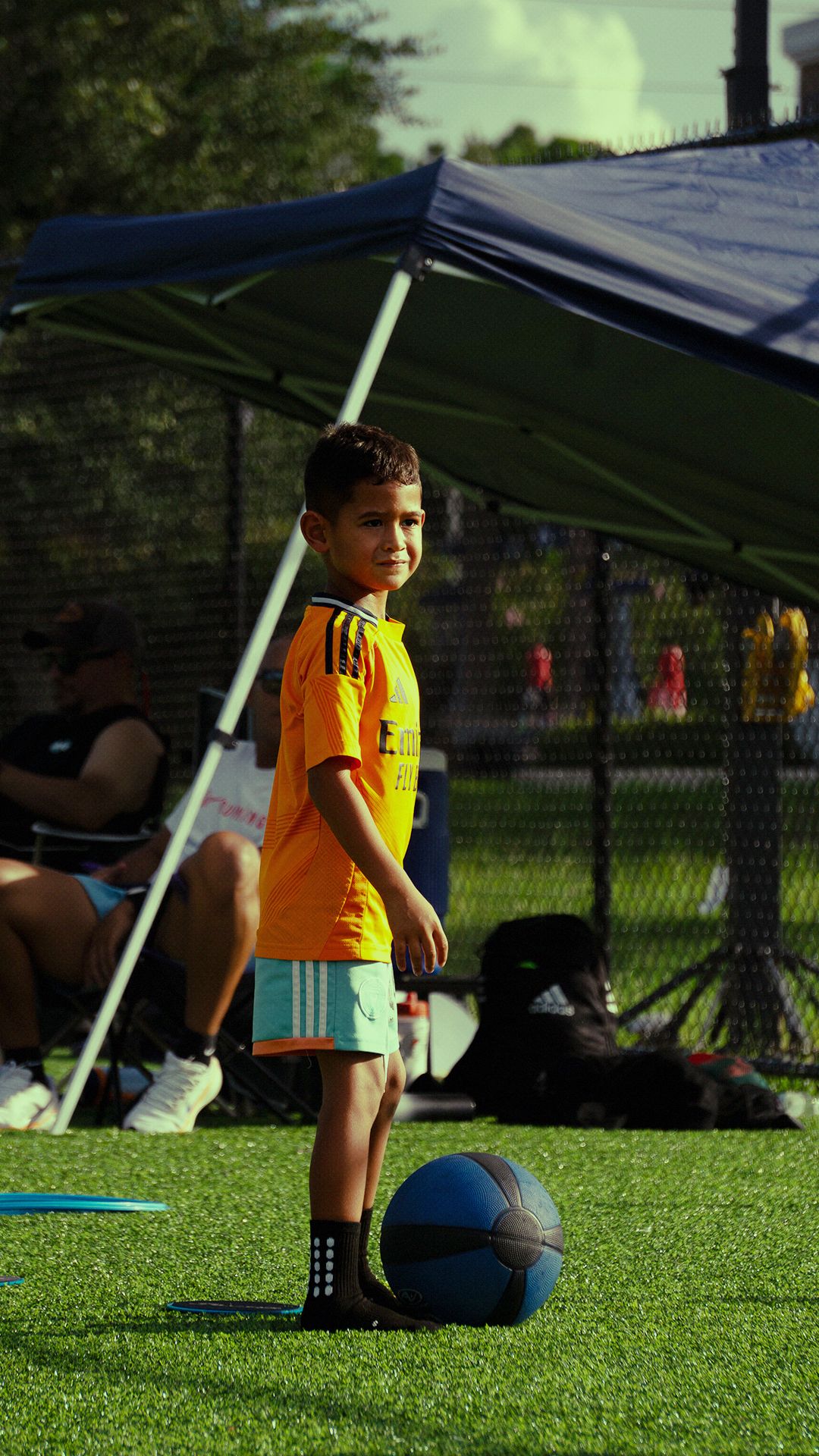 Young boy in yellow Adidas soccer jersey stands beside a blue medicine ball on a green turf field.