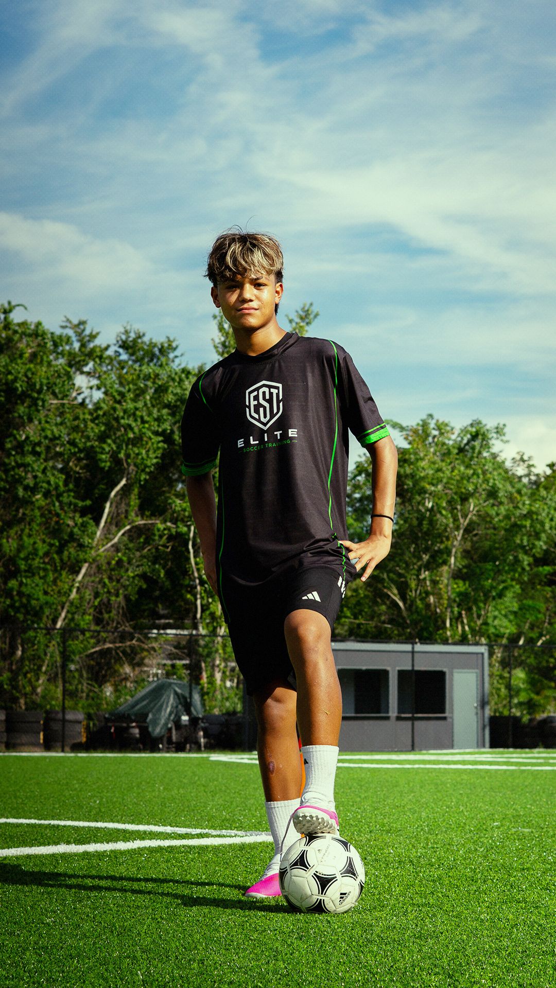 Teen soccer player in black Elite Soccer Training jersey poses with ball on turf field.