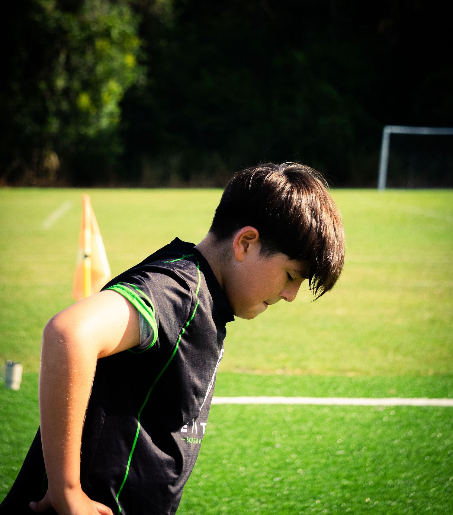 Young boy in black and green jersey bending forward on a green soccer training field with orange cones.