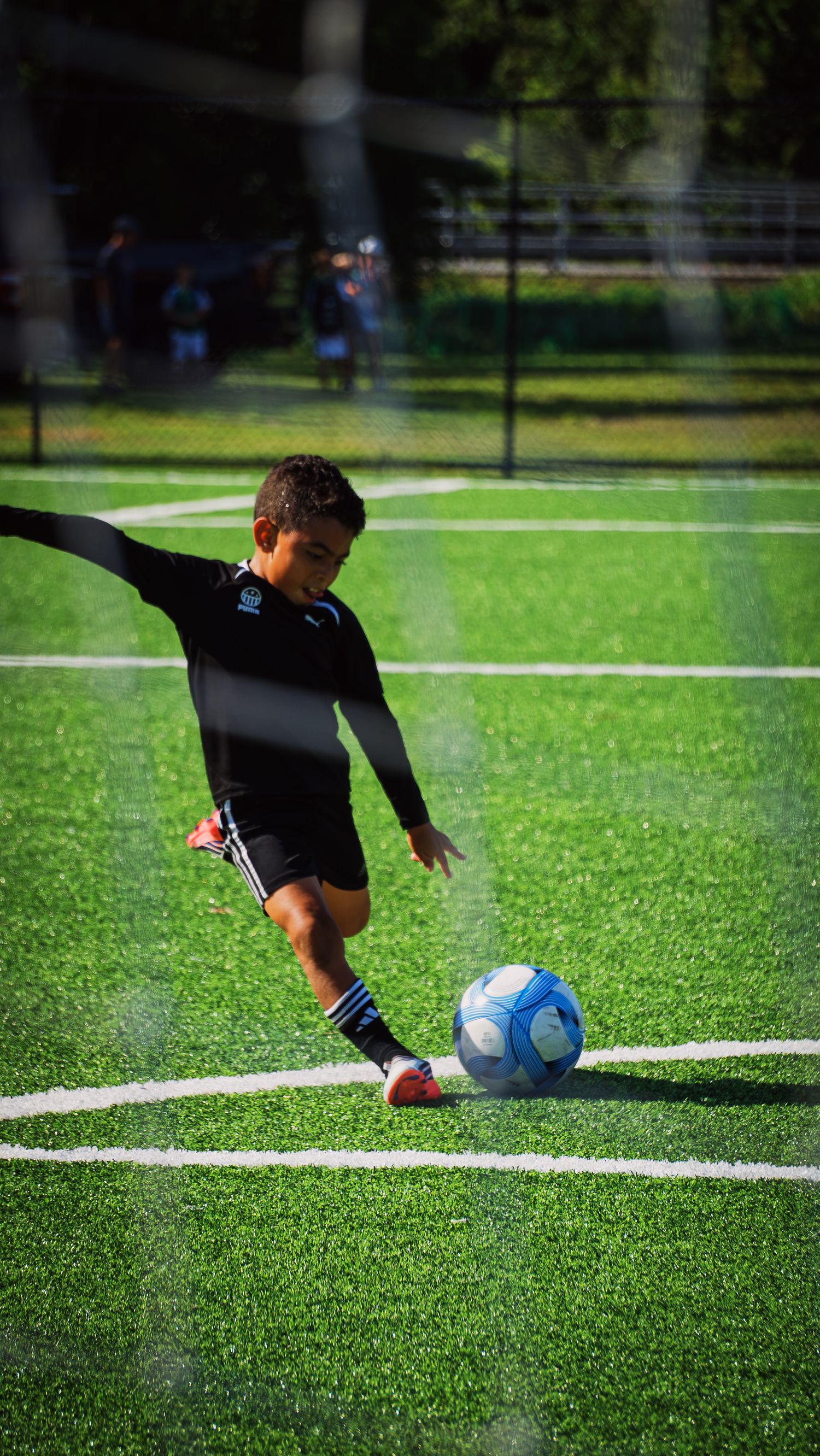 Young boy in black kit striking a blue soccer ball on bright green artificial turf field.