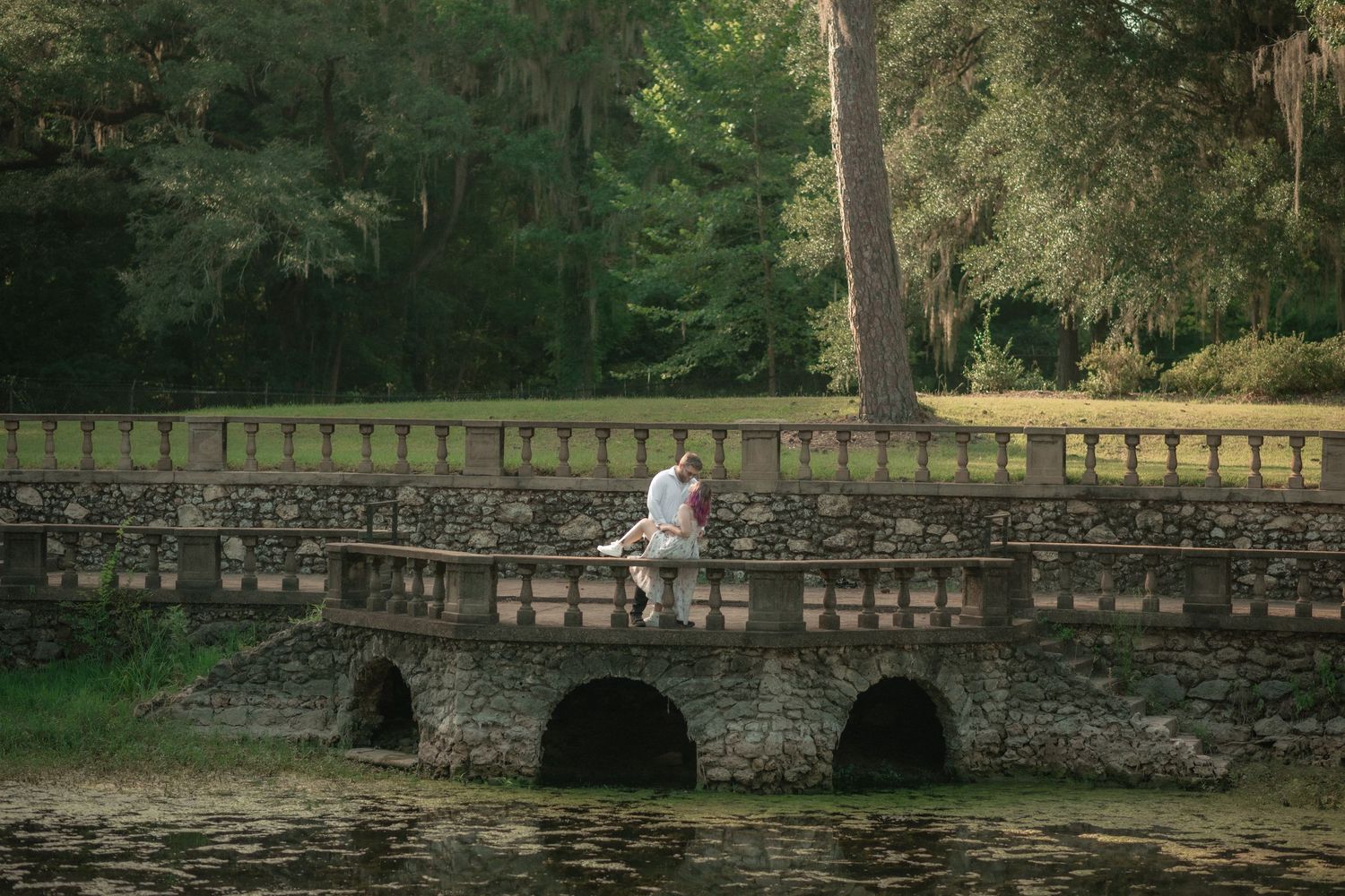 Wedding couple sits together on a stone bridge arch over water surrounded by lush green trees.