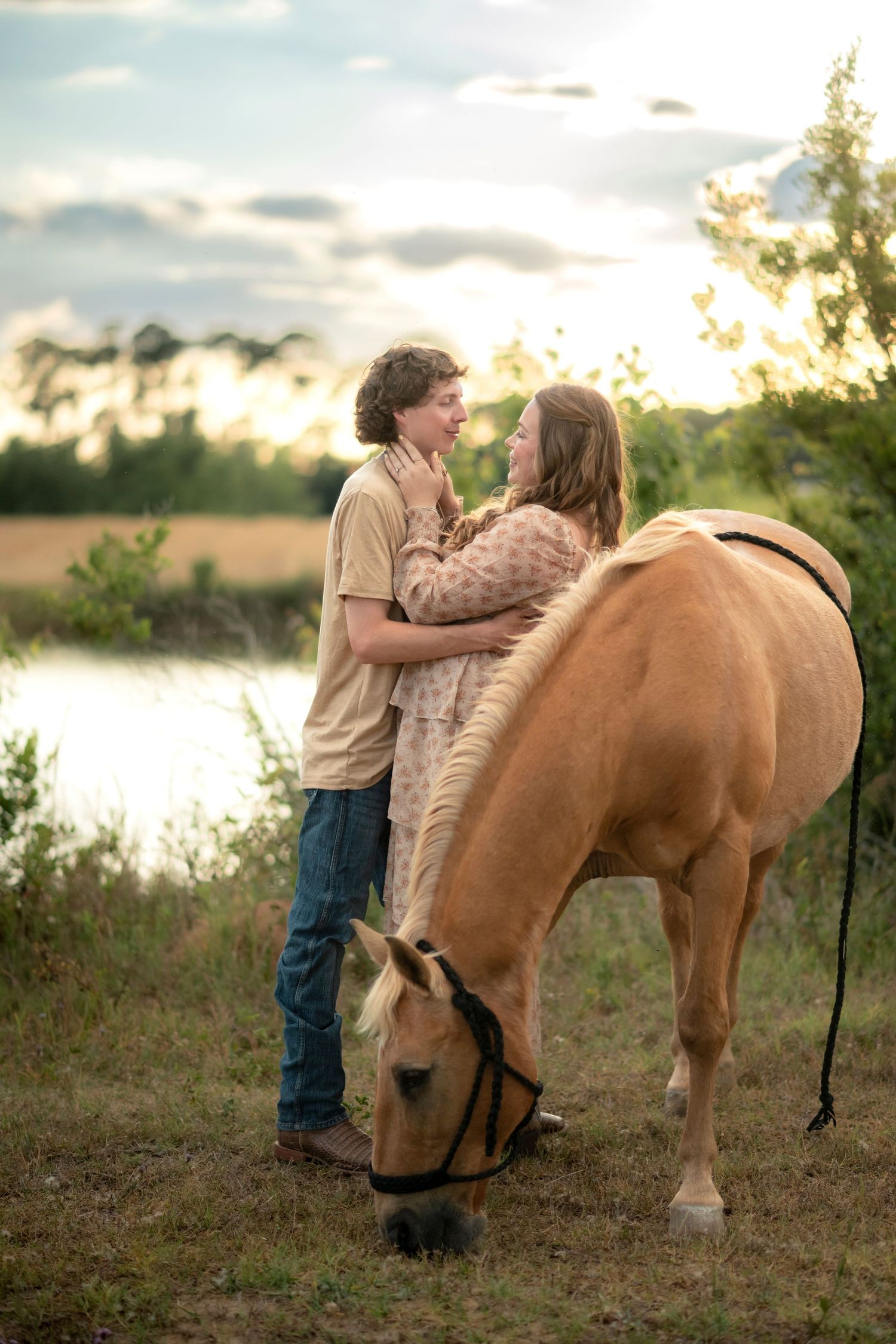 Young romantic couple embraces near a grazing palomino horse by a lake at golden sunset.