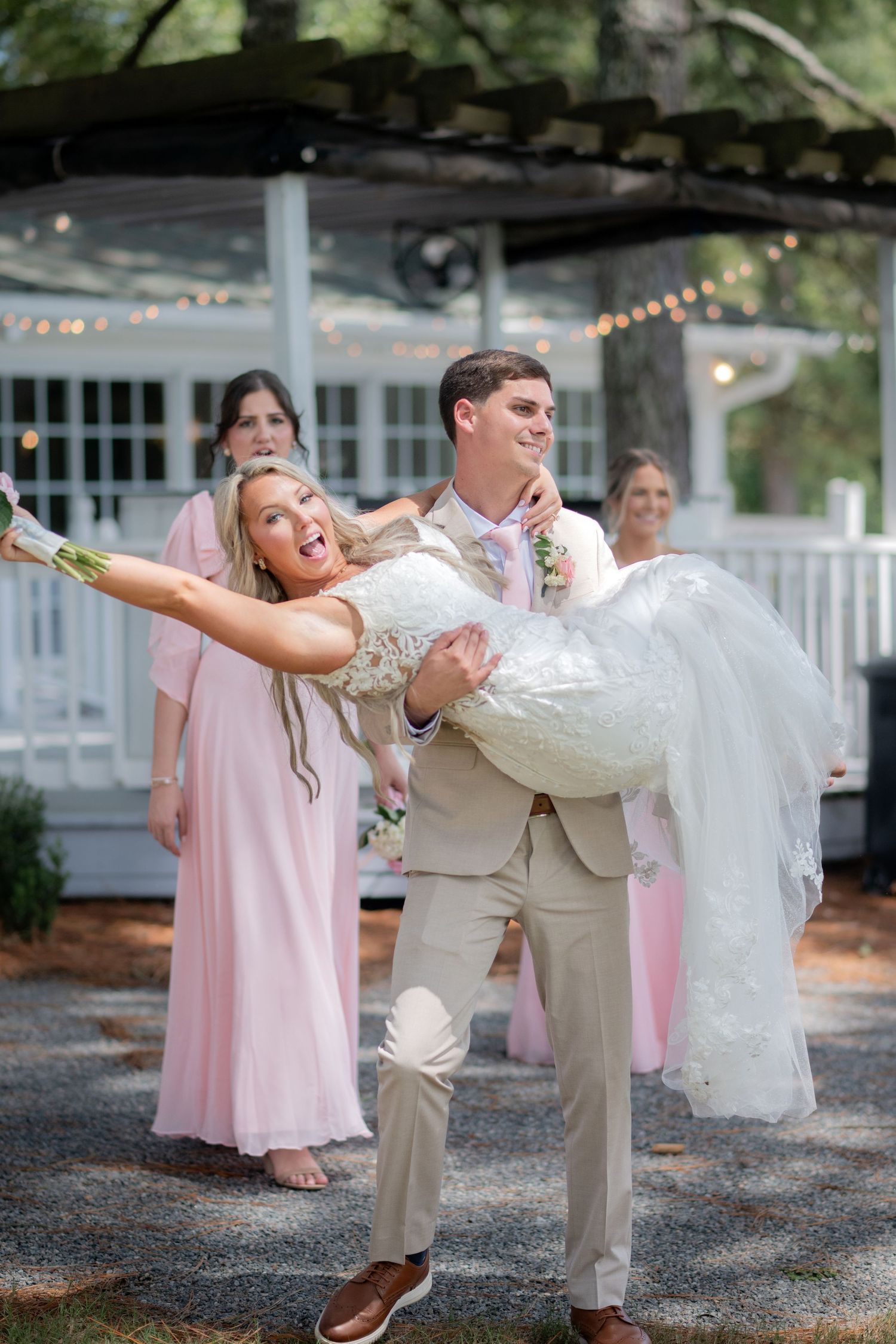 Excited bride being lifted by groom in tan suit while bridesmaids in pink dresses look on joyfully.