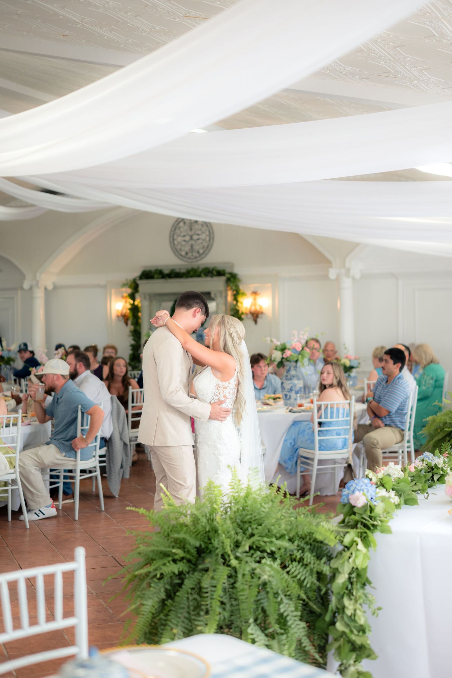 Couple shares first dance at elegant wedding reception with white draped ceiling, fern centerpieces, and guests seated at decorated tables.