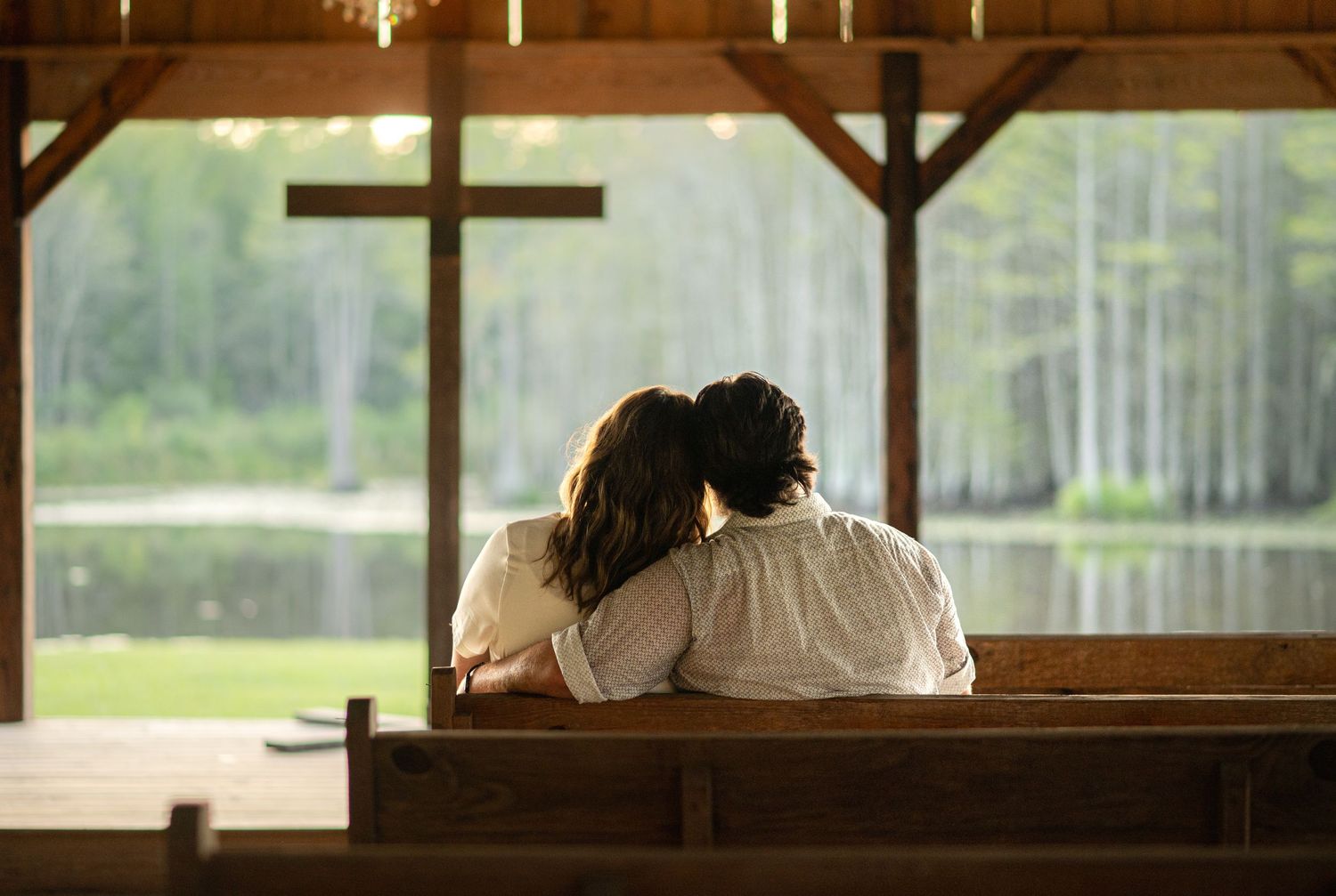 Couple sitting closely together on a wooden church pew in a rustic chapel overlooking a serene lake.