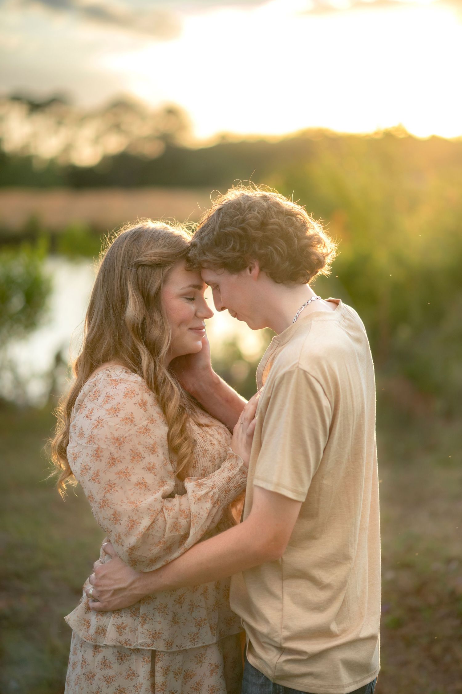 Young couple embraces forehead-to-forehead in a romantic golden sunset glow by a tranquil lake.
