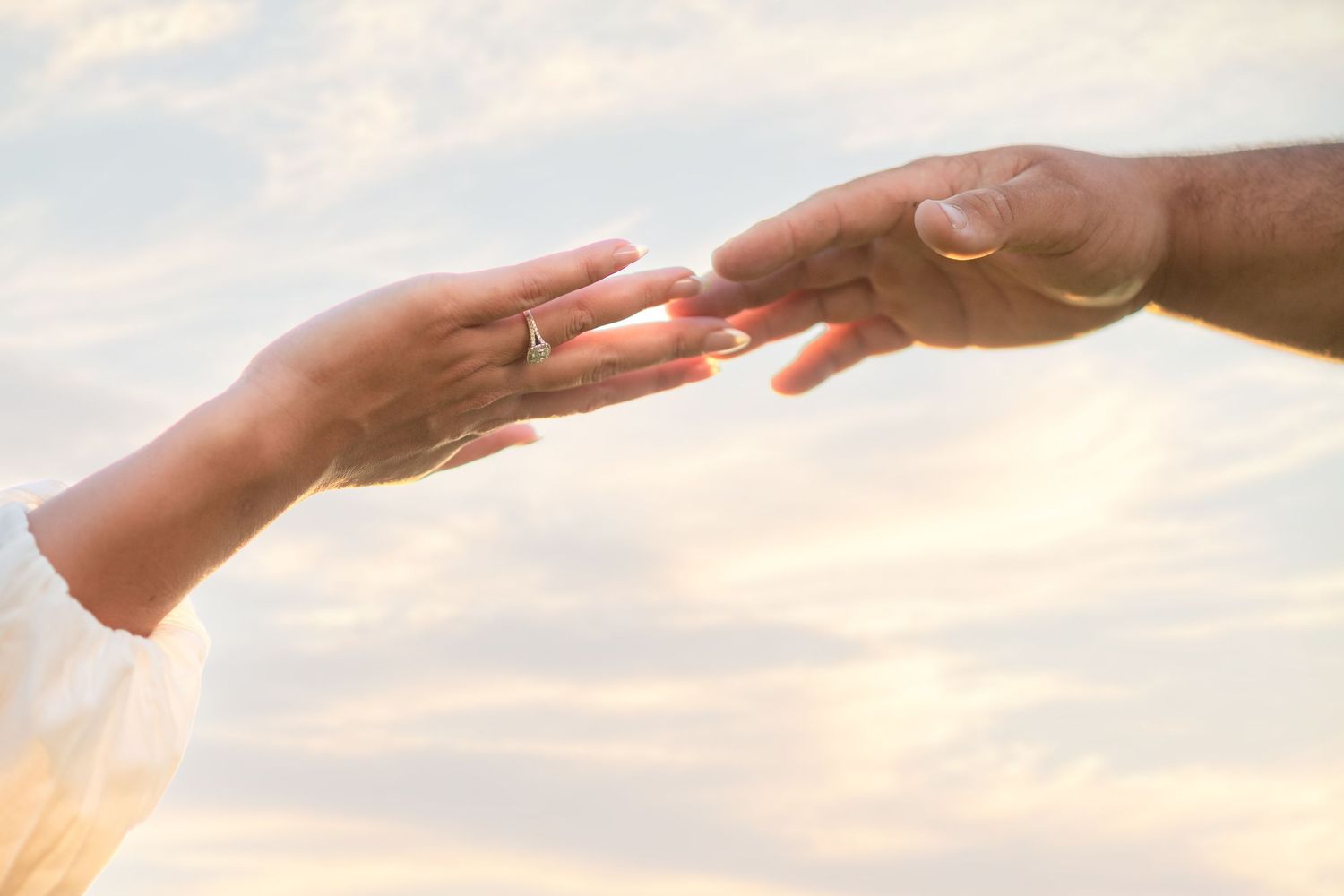 Two hands gently reaching toward each other with a golden engagement ring visible against a cloudy sky.