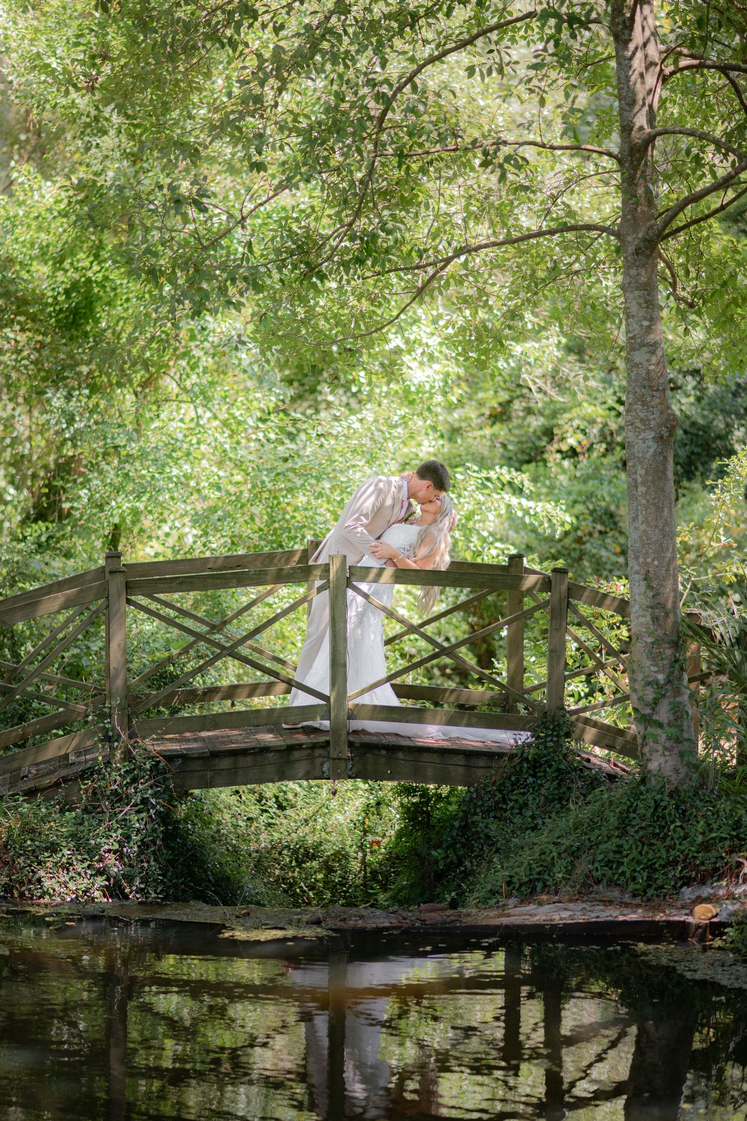 Wedding couple sharing a romantic kiss on a rustic wooden bridge over a pond in a lush green forest.