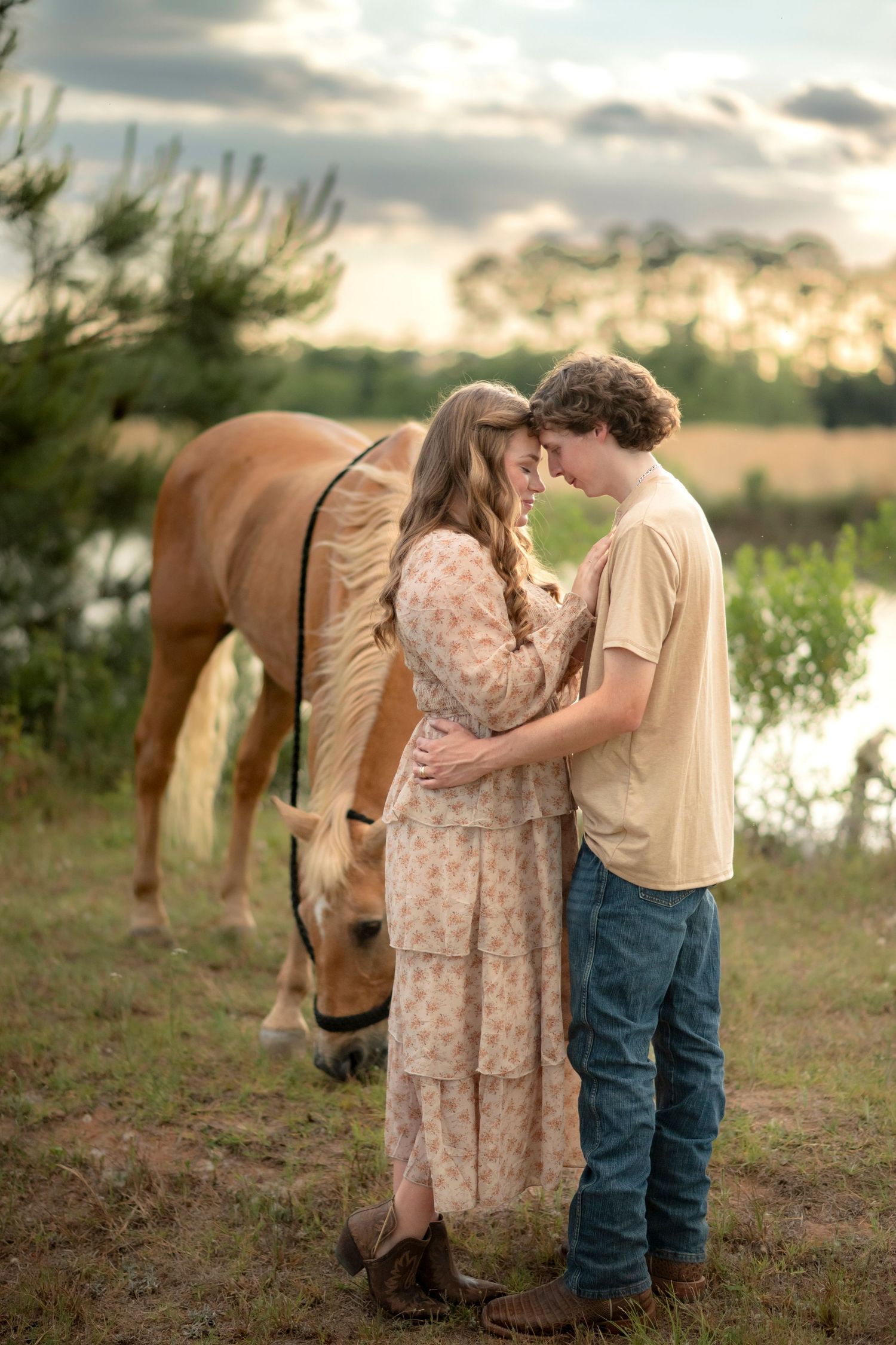 Couple shares a tender moment together beside a grazing palomino horse at a scenic lakeside sunset.