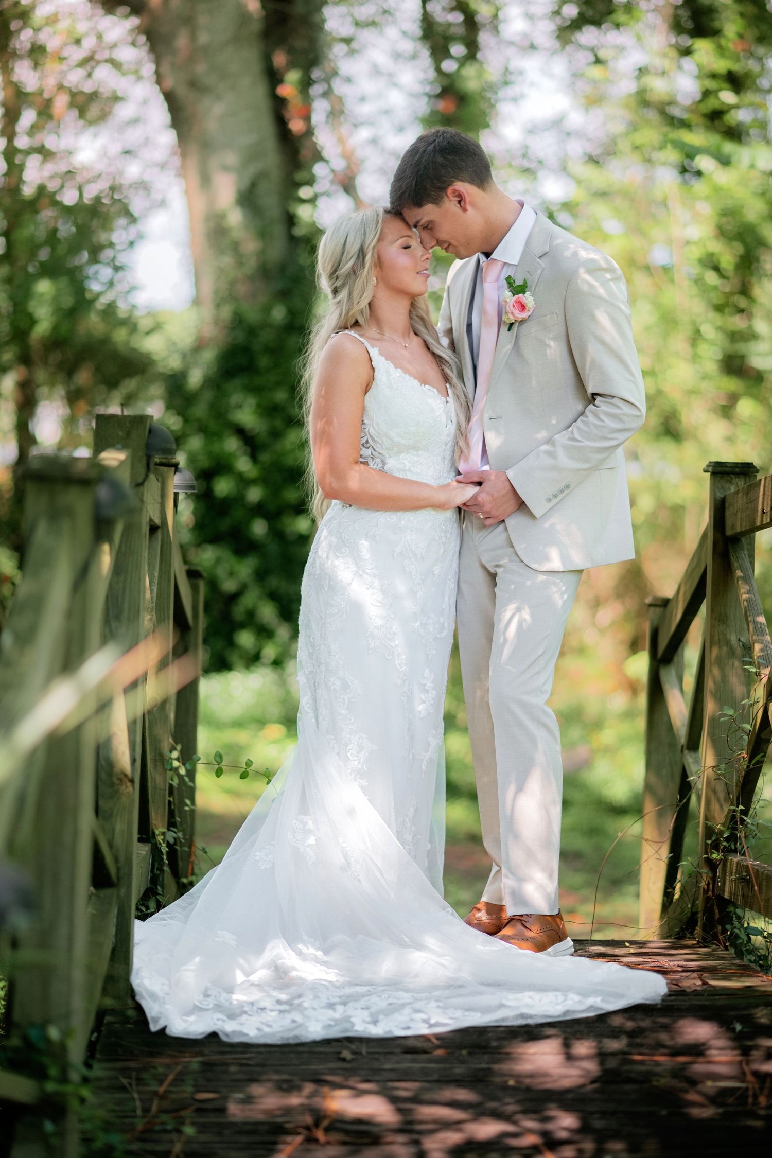 Bride in elegant lace gown and groom in tan suit share a tender moment on a sunlit garden bridge.