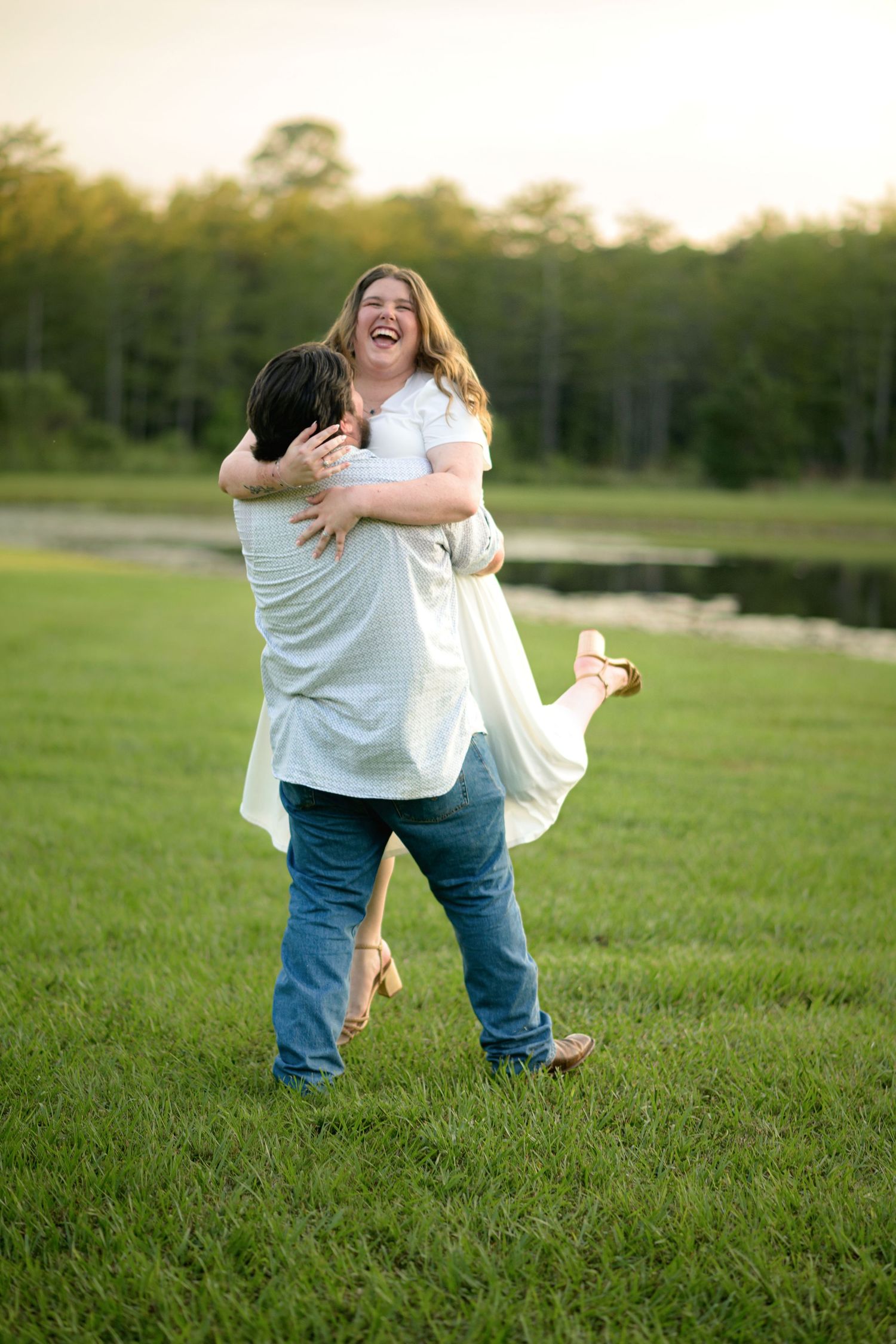 Joyful couple sharing a romantic moment as man lifts laughing woman in a lush green field at sunset.