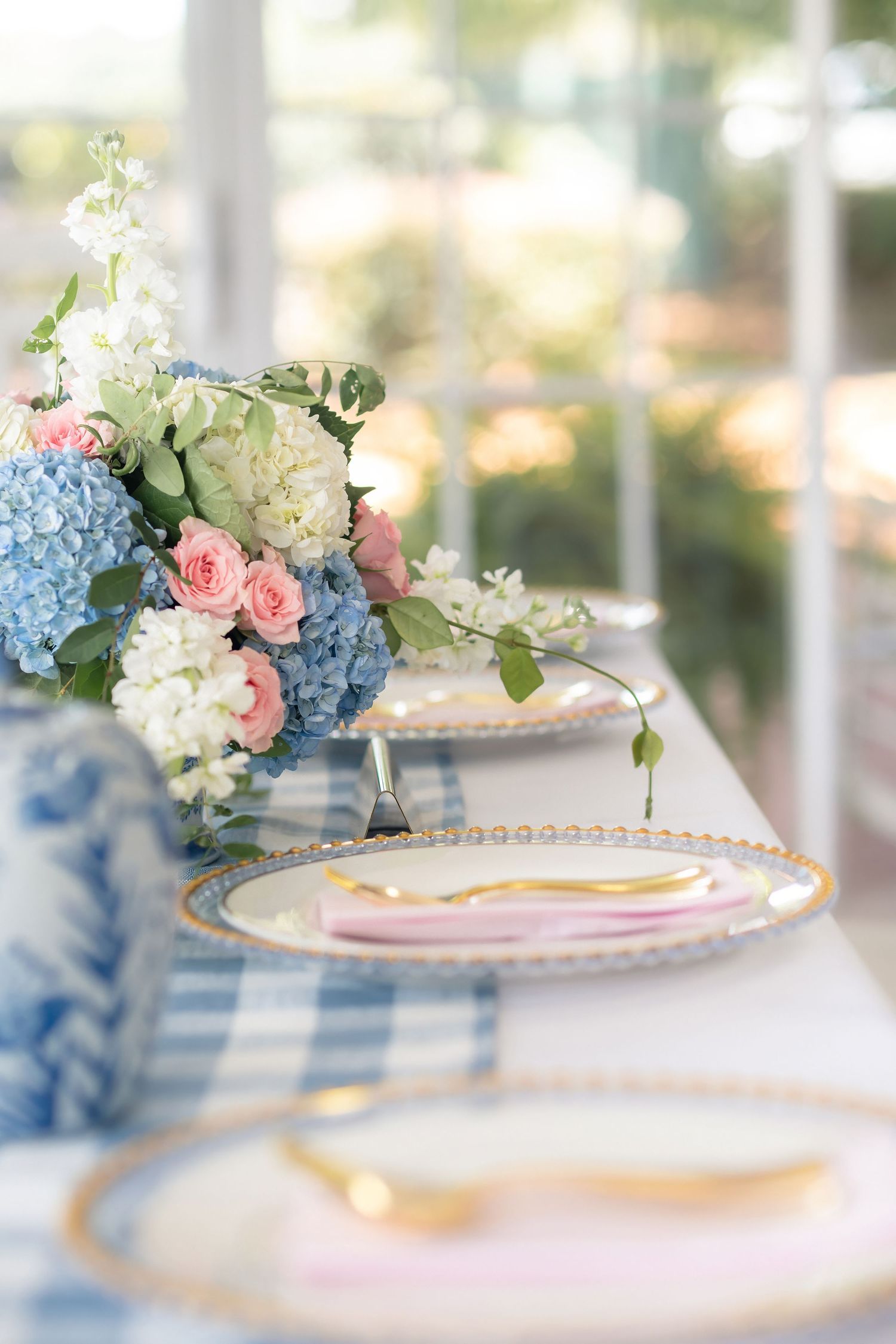 Elegant wedding table setting with gold-rimmed plates, pink napkins, and a blue hydrangea and pink rose floral centerpiece.