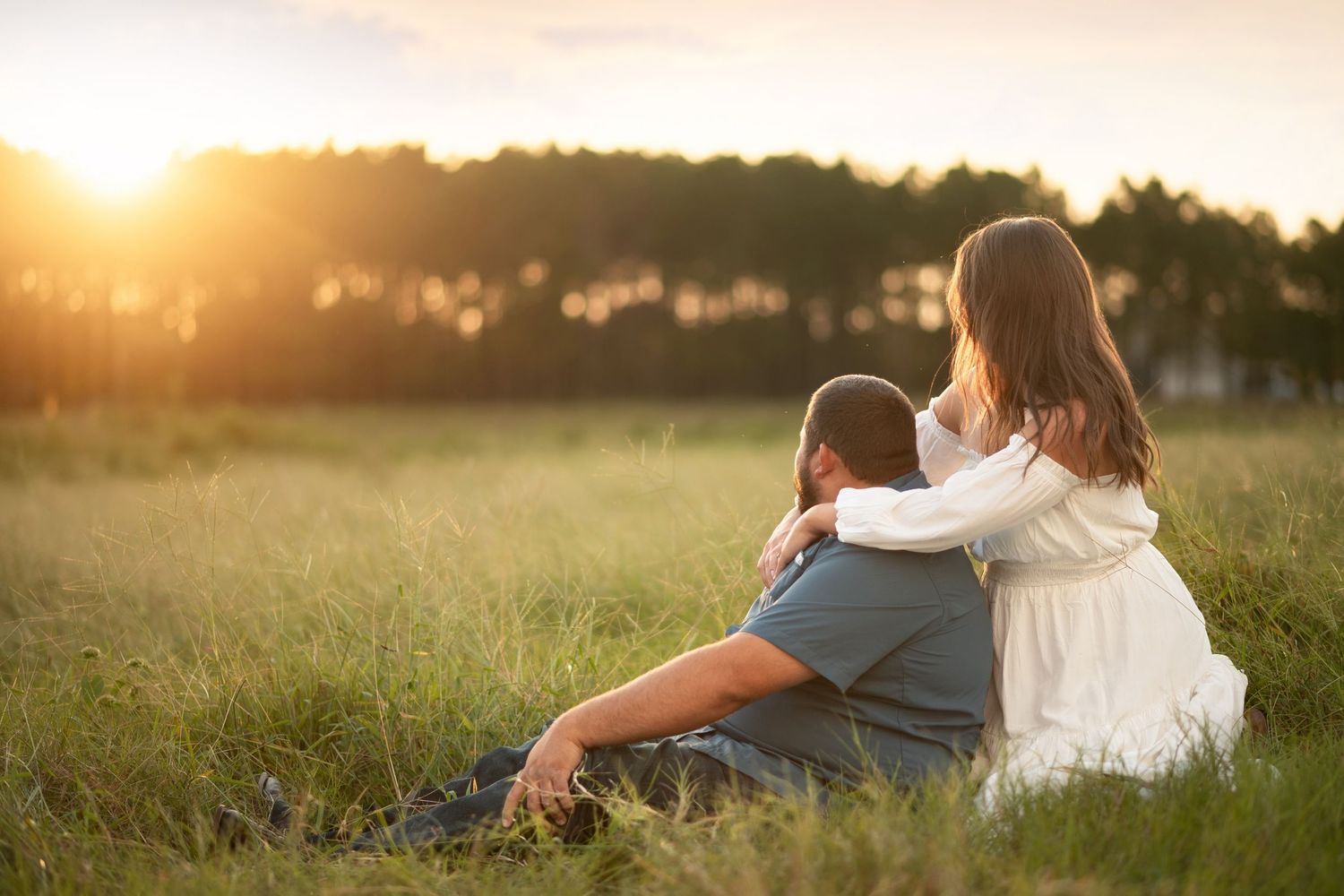 Couple sitting together in a golden meadow at sunset, woman embracing man from behind in peaceful moment.