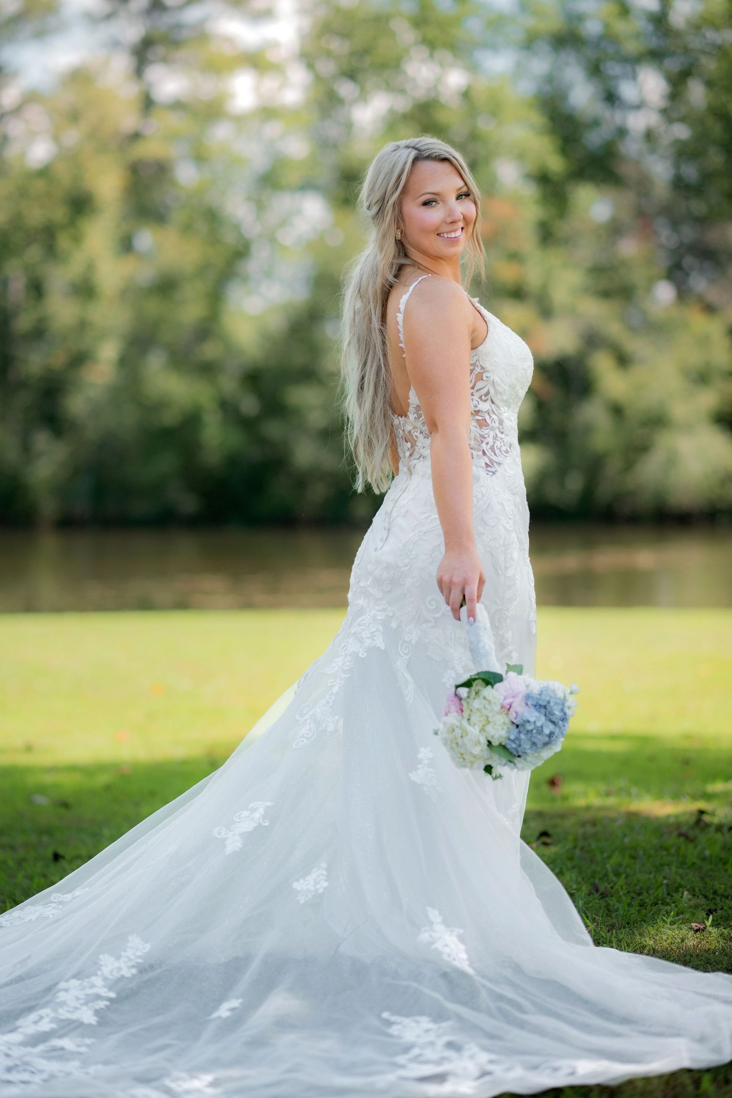 Radiant blonde bride in lace mermaid gown smiles while holding a blue hydrangea bouquet near a lake.