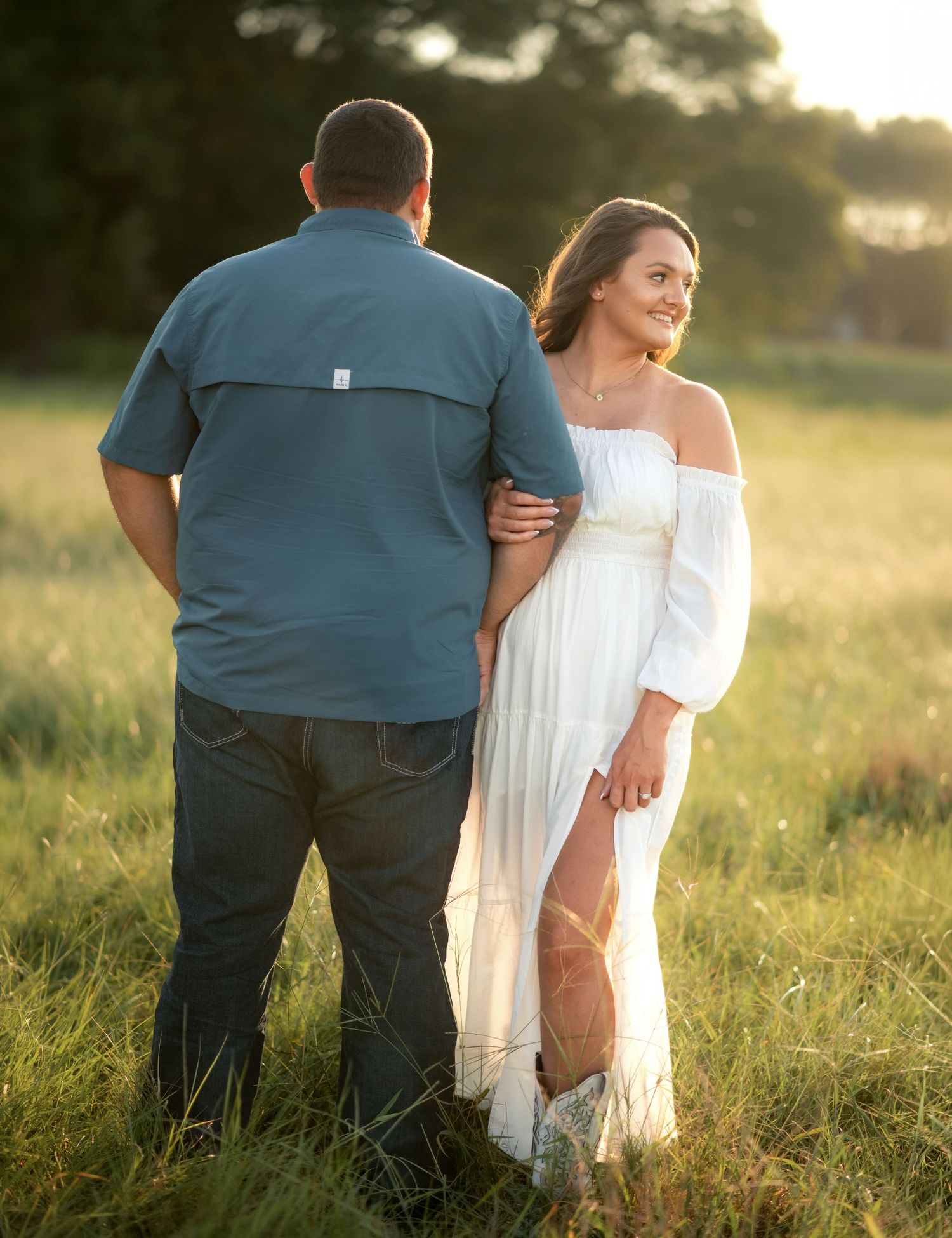 Couple standing in a golden field at sunset, woman in white dress smiling while embracing her partner.