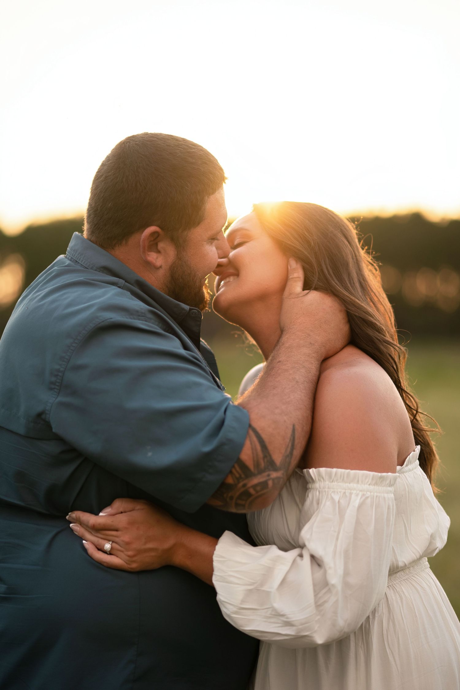 Couple sharing a romantic kiss at golden hour sunset in an outdoor field, man with tattoos embracing woman in white dress.
