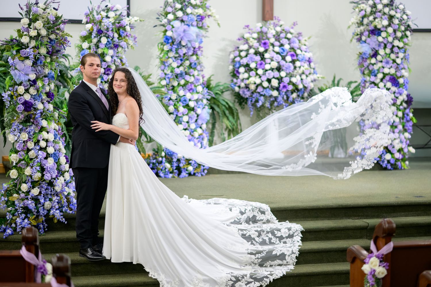 Wedding couple poses at church altar surrounded by stunning blue and white floral arrangements, veil flowing.