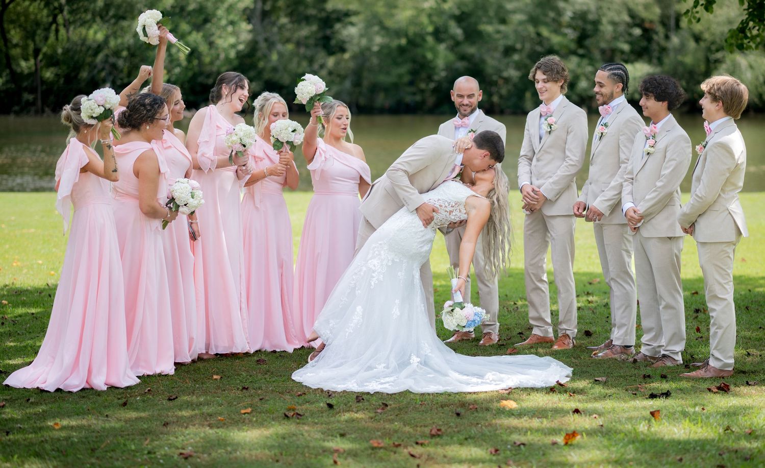 Bride and groom share a romantic dip kiss surrounded by wedding party in pink and tan attire outdoors.