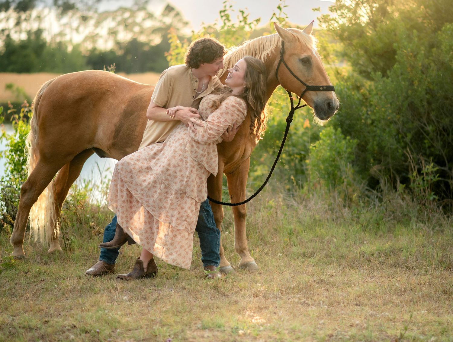 Romantic couple sharing a kiss beside a golden horse in a sunlit meadow during an engagement shoot.