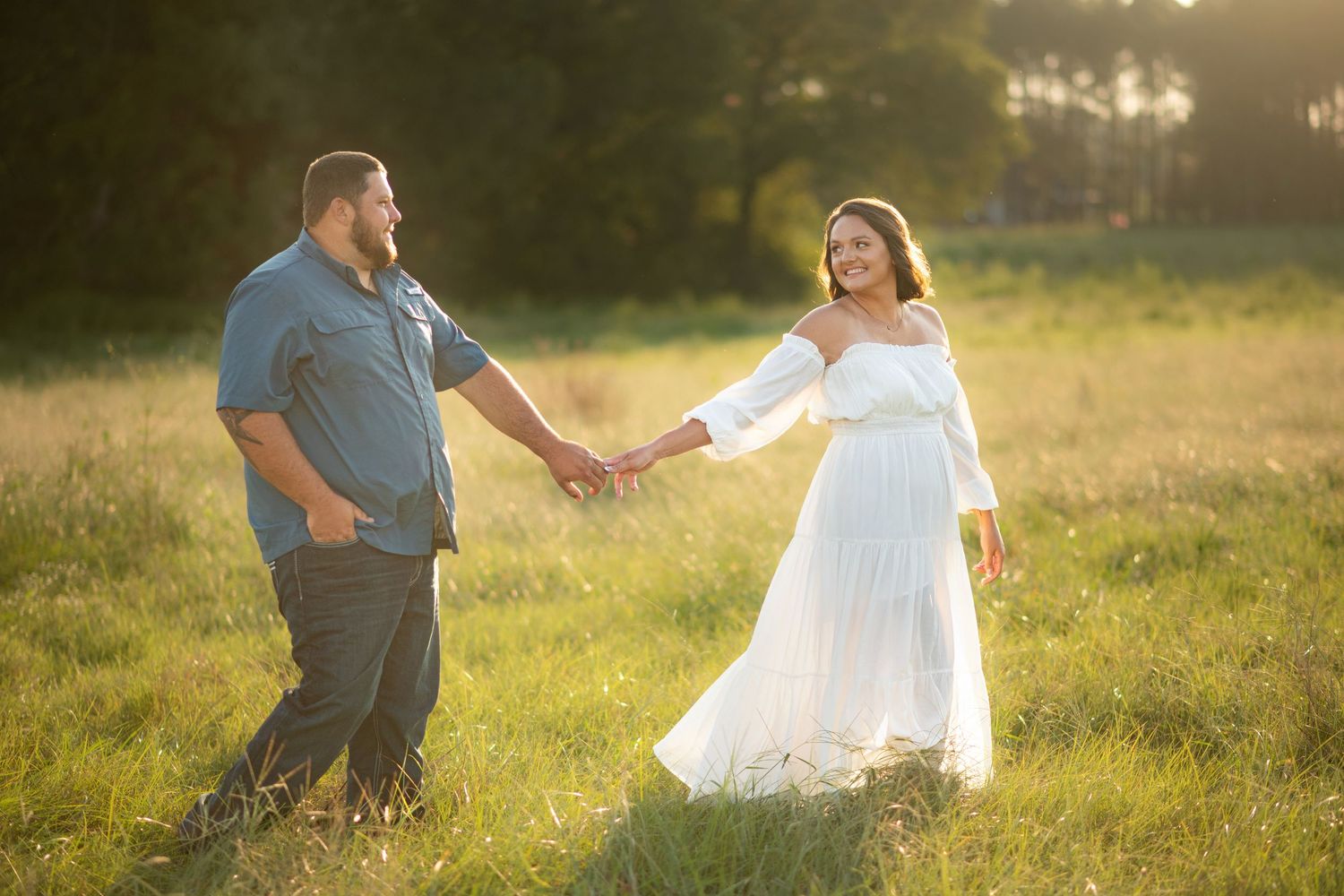 Engaged couple holding hands and walking through a sunlit meadow, woman wearing a flowing white dress.