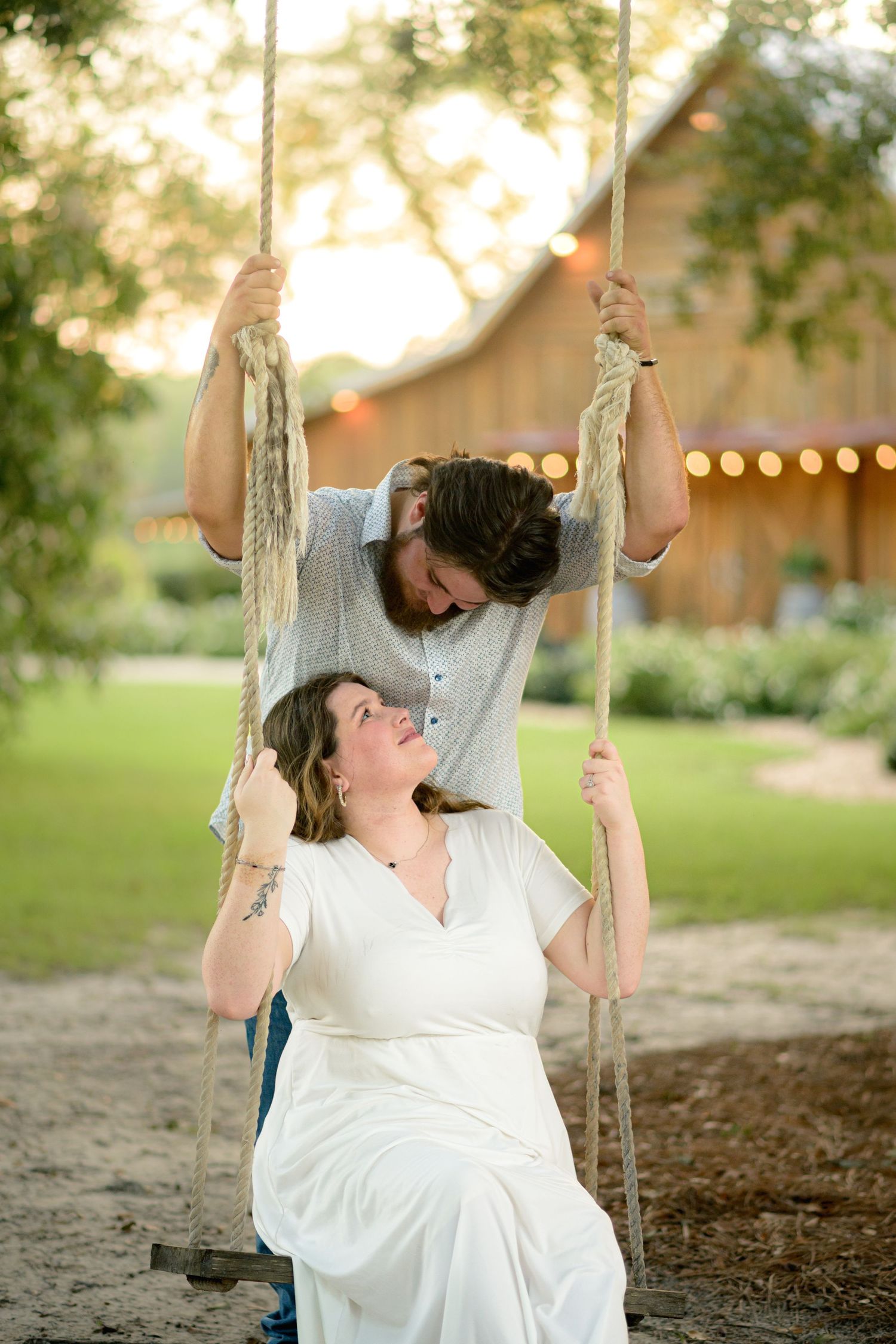 Man lovingly looks down at woman sitting on a rope swing at a rustic barn venue.