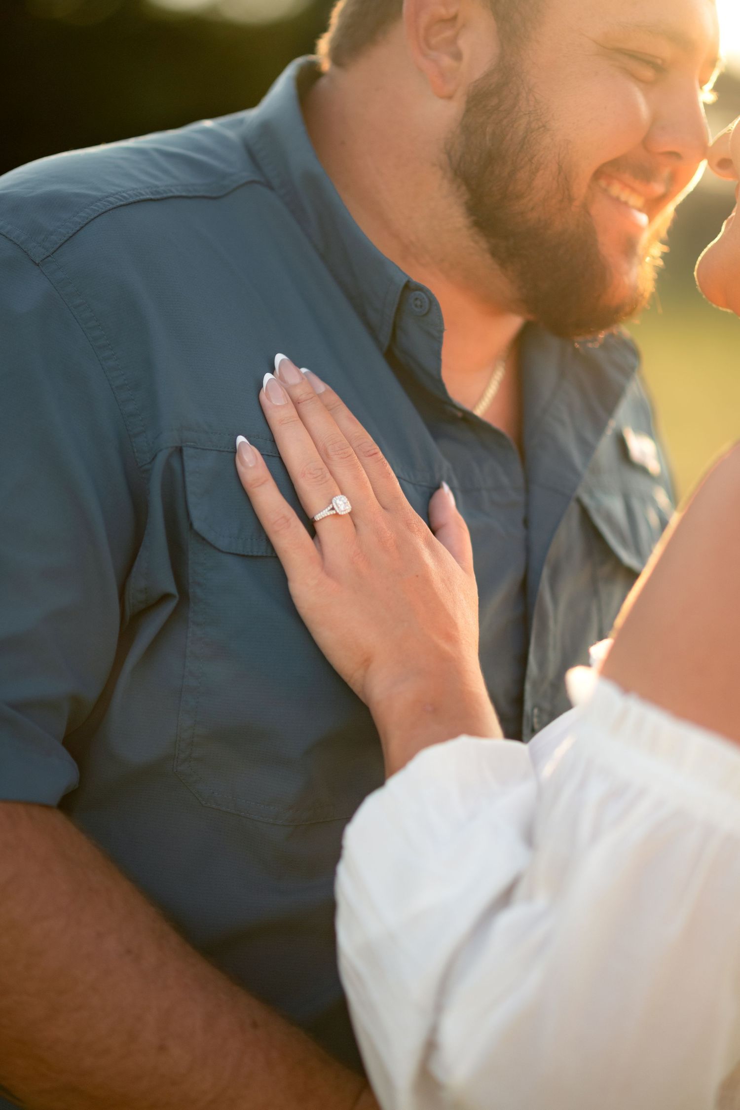 Close-up of woman showing off her diamond engagement ring against her smiling bearded partner's blue shirt.