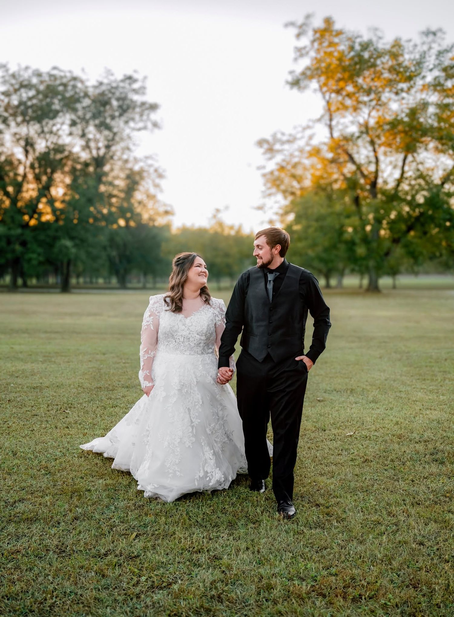 Bride and groom walk hand in hand through a green park at golden hour in their wedding attire.