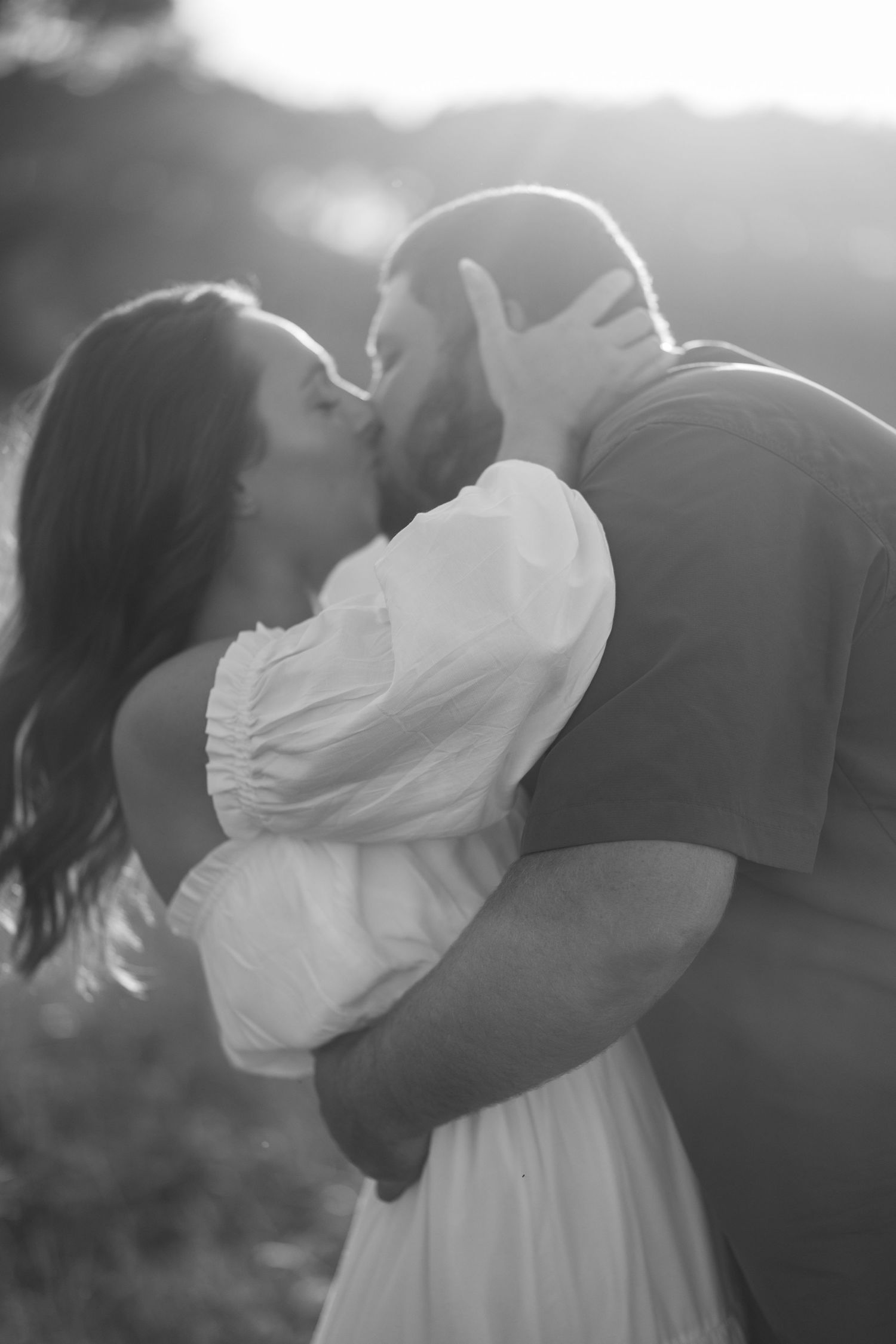 Black and white photo of a couple sharing a passionate kiss in a sun-drenched outdoor setting.