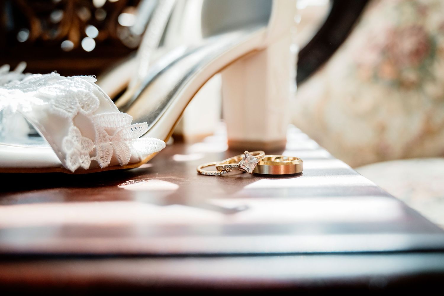 Wedding details flat lay featuring bridal heels with lace garter and gold wedding rings on a wooden table.