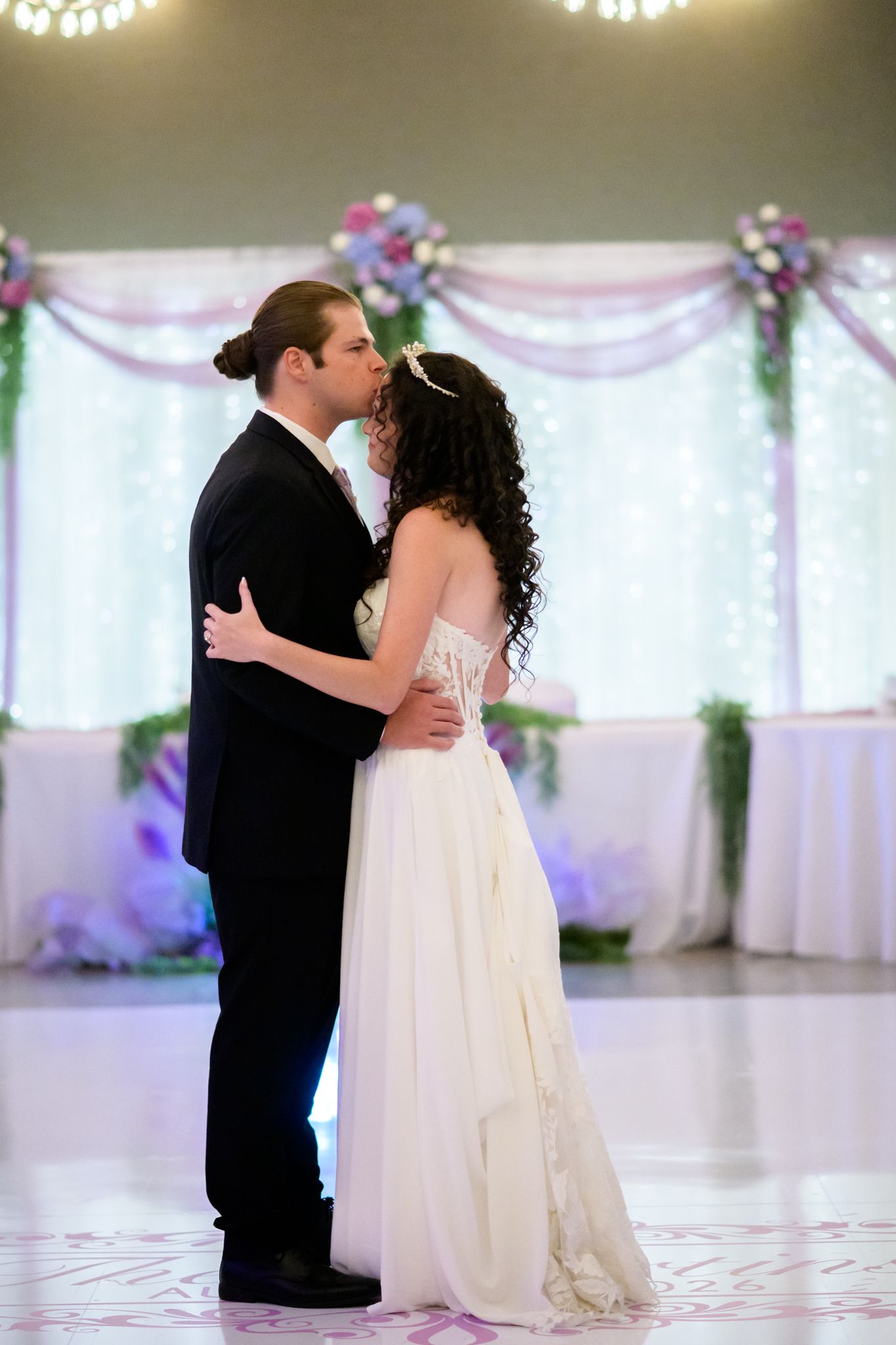 Bride and groom share their first dance kiss in an elegantly decorated ballroom with floral arrangements.