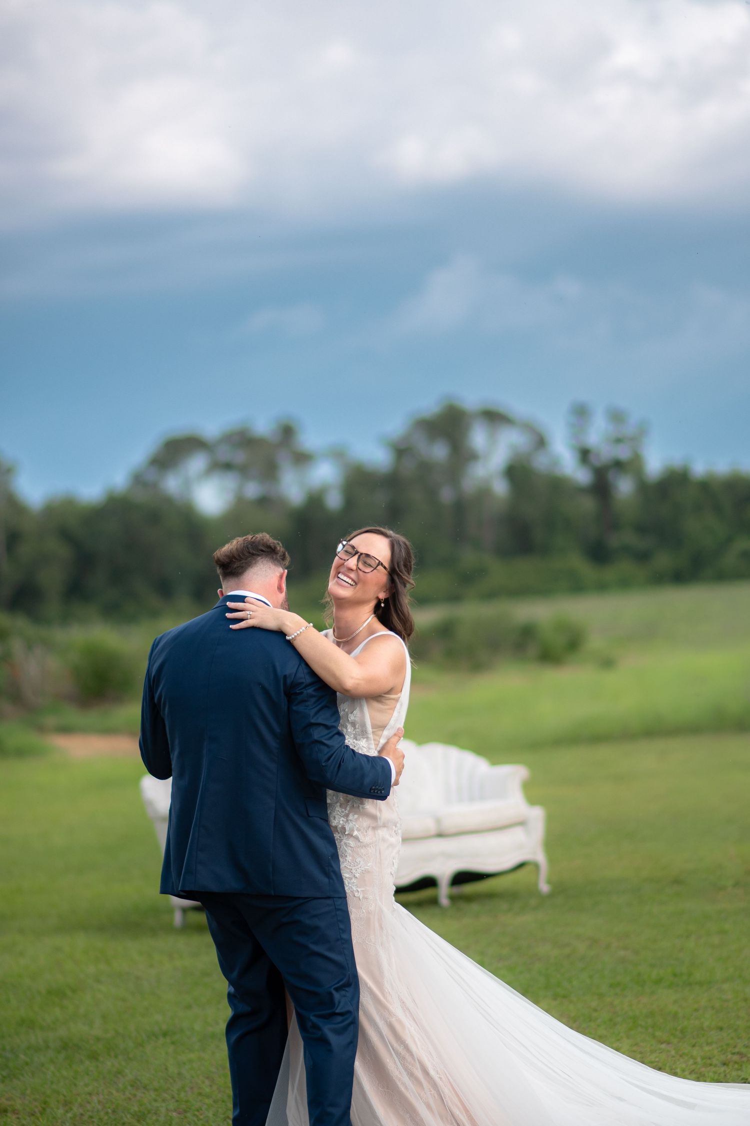 Smiling bride in white gown embraces groom in navy suit on a lush green outdoor lawn.