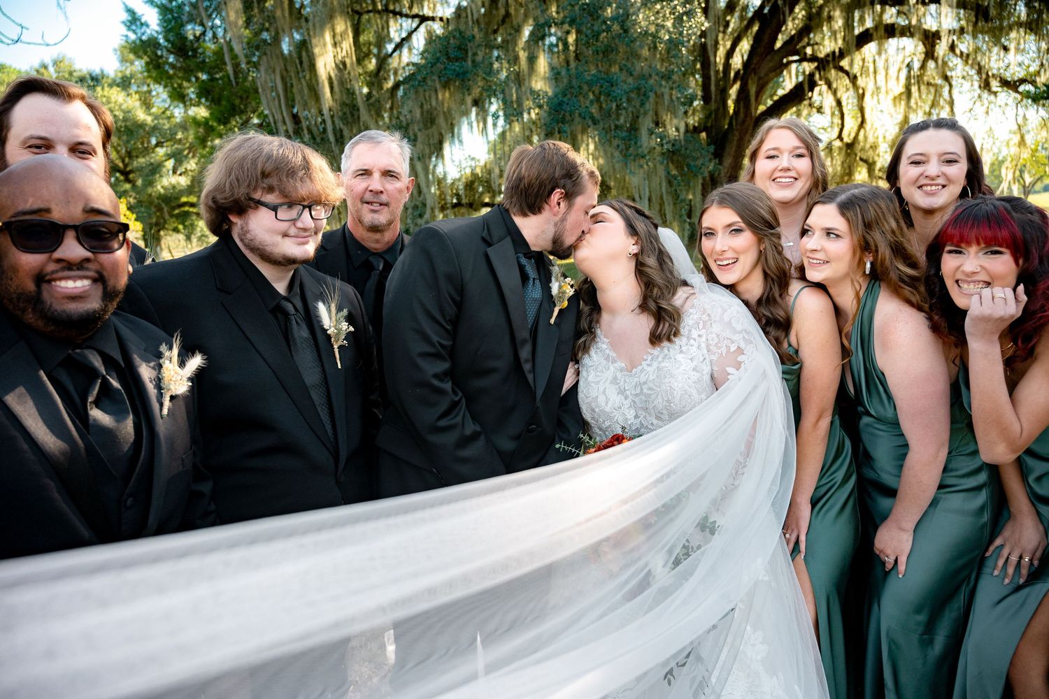 Wedding party smiling as bride and groom share a kiss outdoors under moss-draped trees.