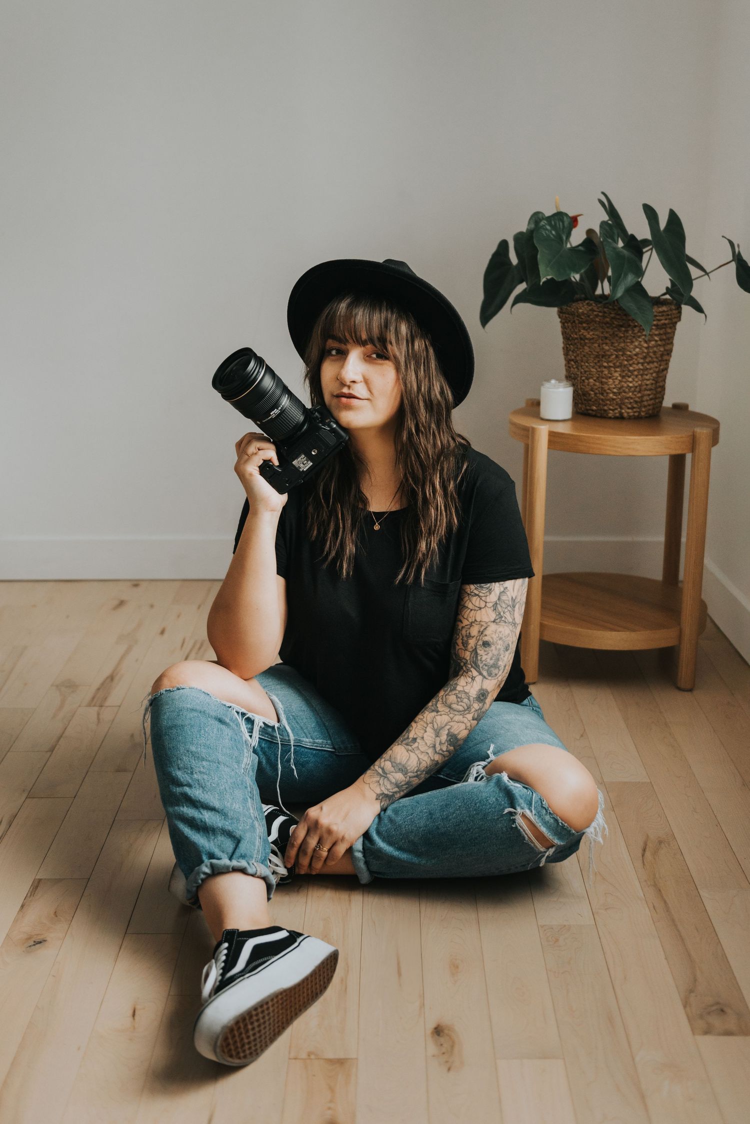 Photographer with tattoos sits cross-legged on wooden floor wearing black hat, holding camera near potted plant.