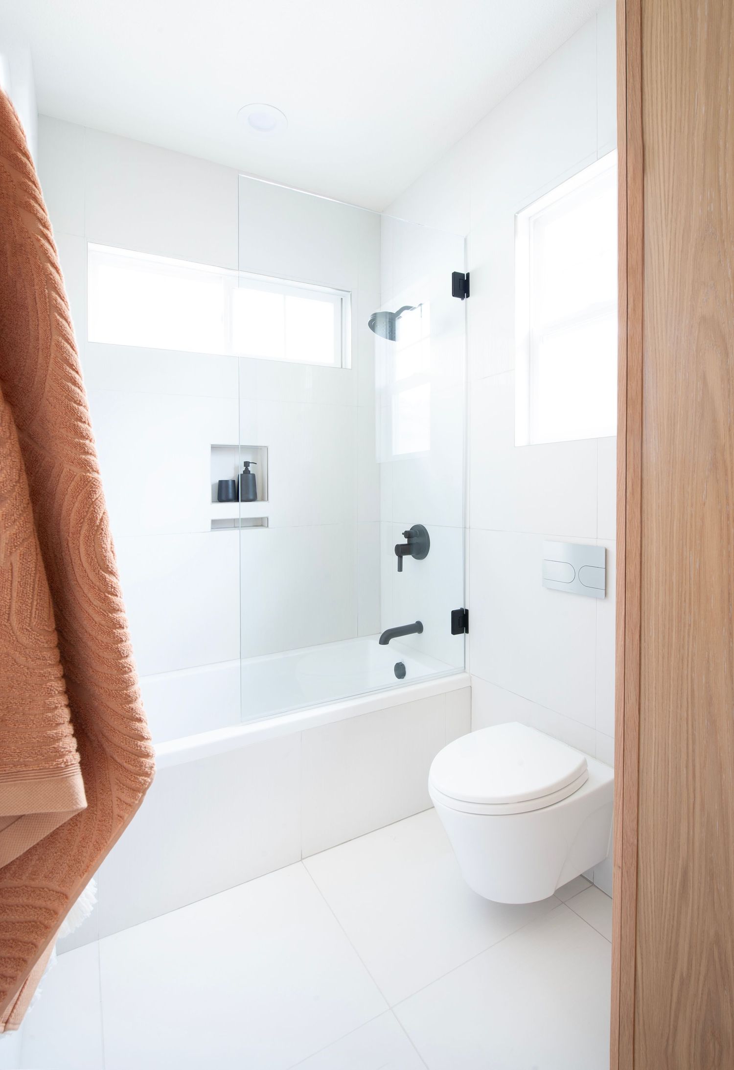 Clean white bathroom featuring a wall-mounted toilet, glass shower enclosure, and terracotta towel accent.