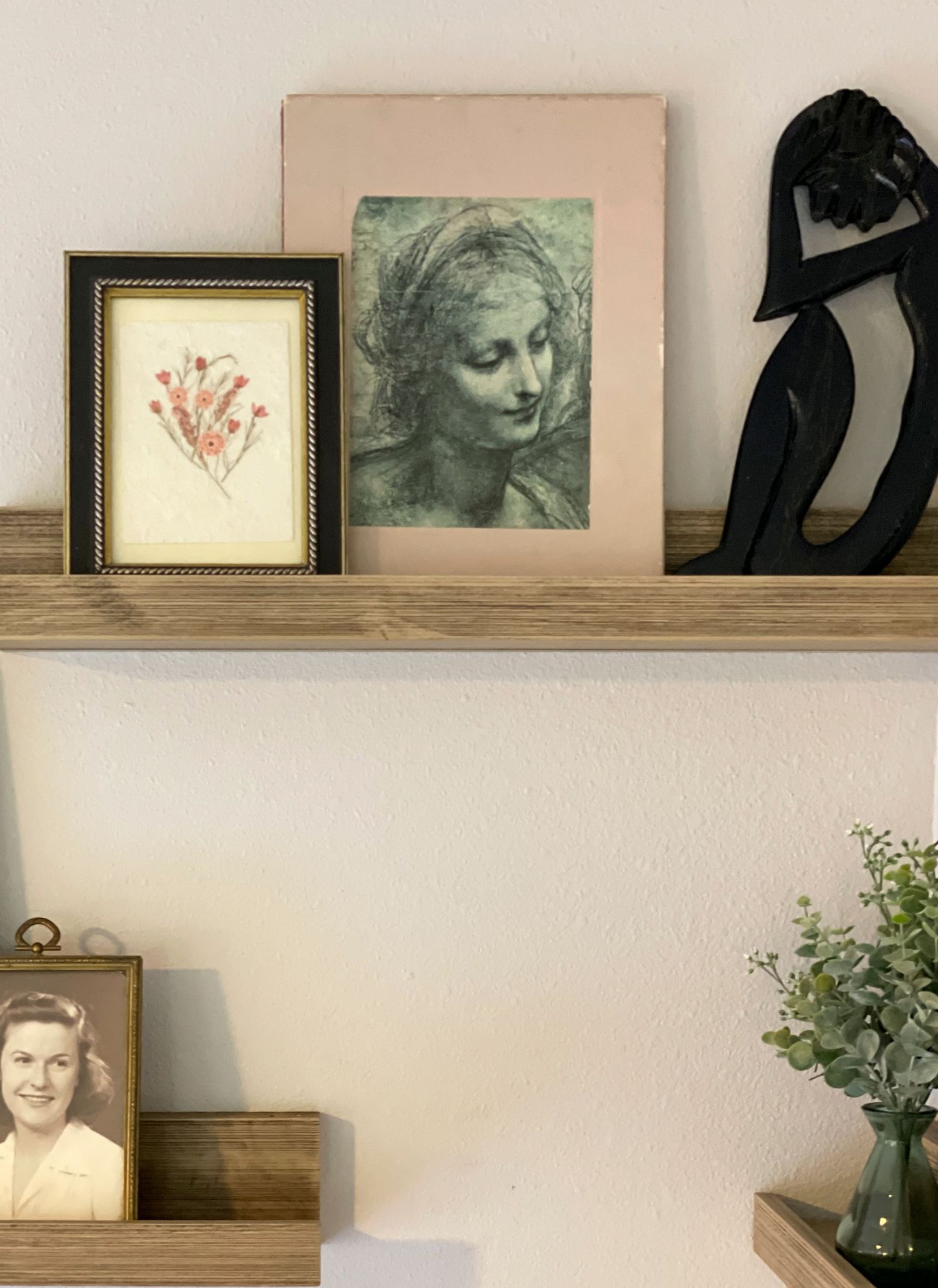 Wooden shelf displaying vintage artwork, decorative frames, and greenery against white wall.