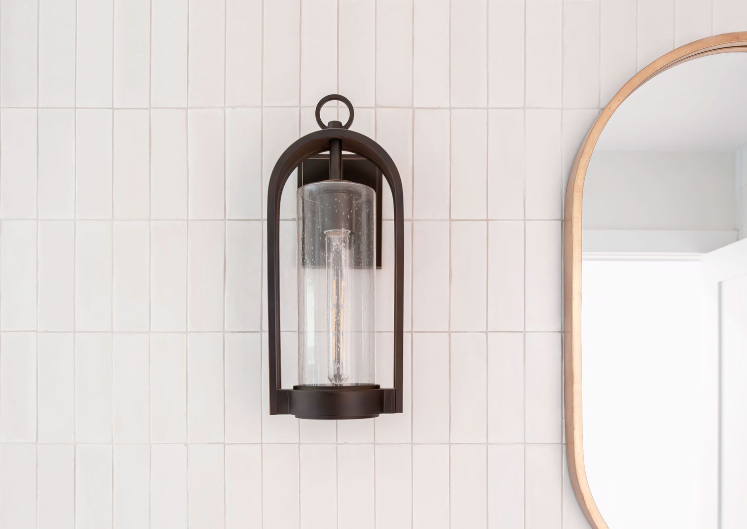 White tiled bathroom wall with black lantern sconce and round wooden mirror.