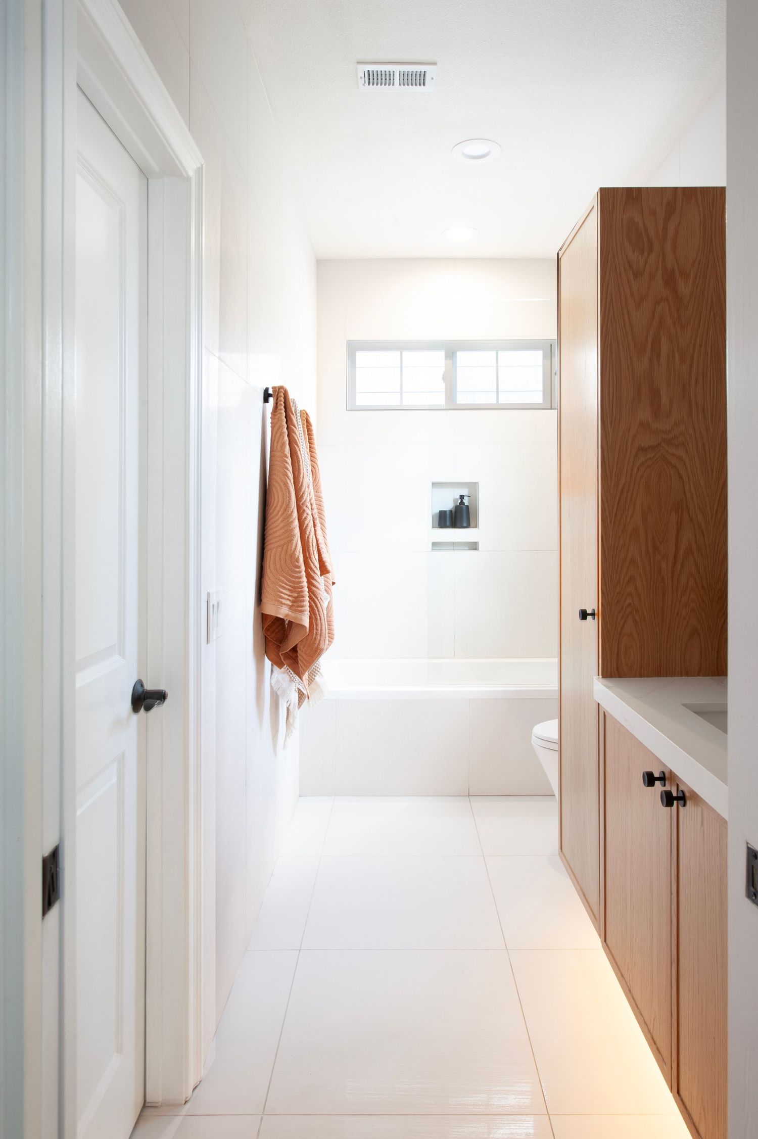 Minimalist white bathroom hallway with wooden storage and warm orange towel accent.