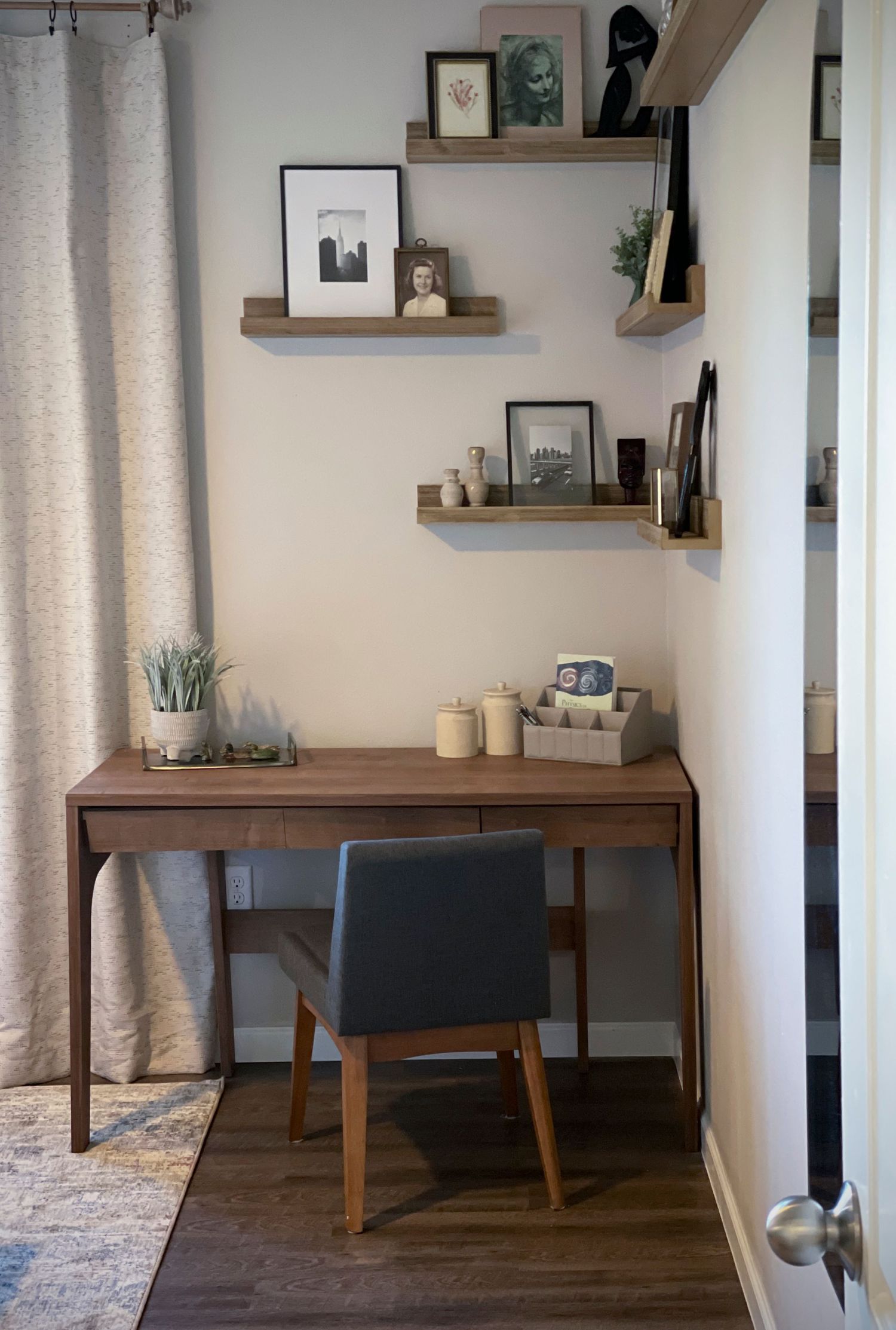 Modern home office setup with wooden desk, grey chair, and floating shelves displaying artwork.