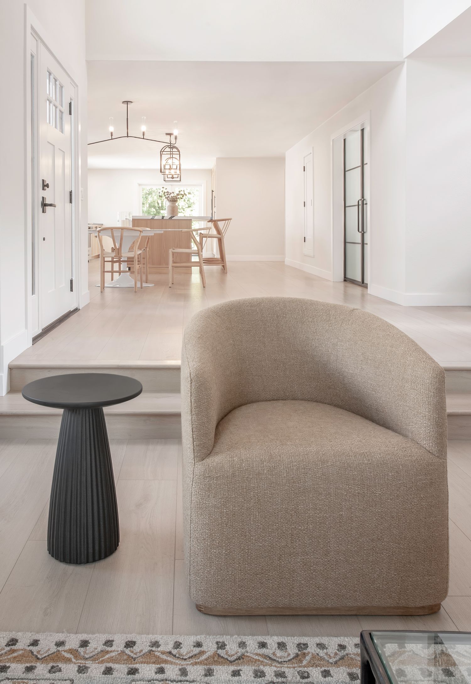 Modern living area featuring curved beige armchair and black pedestal side table on patterned rug in foreground with modern kitchen in back.