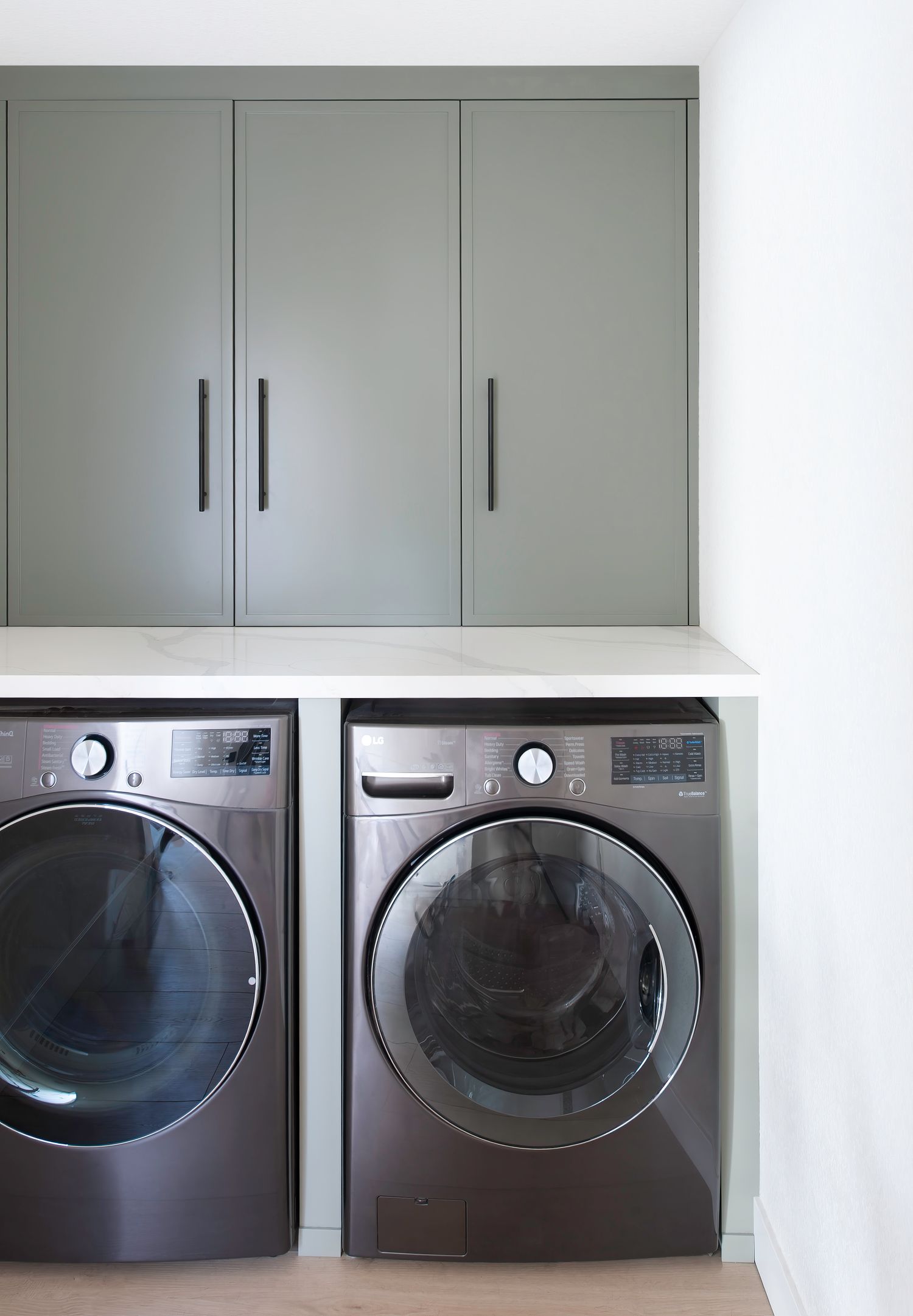 Laundry room with soft gray/green upper cabinets and modern front-loading washer and dryer set.